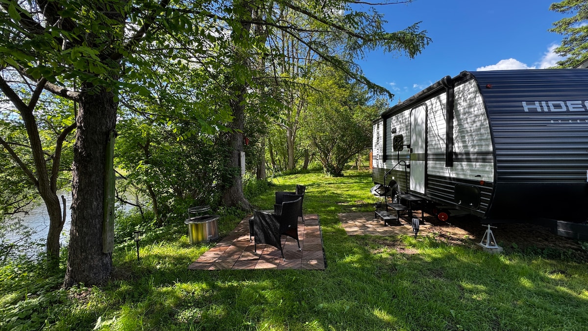 A modern trailer is positioned beside a serene river, surrounded by lush greenery. A cozy outdoor seating area with two chairs is set on a stone patio, while a small fire pit nestles nearby. Sunlight filters through the trees, enhancing the tranquil setting.