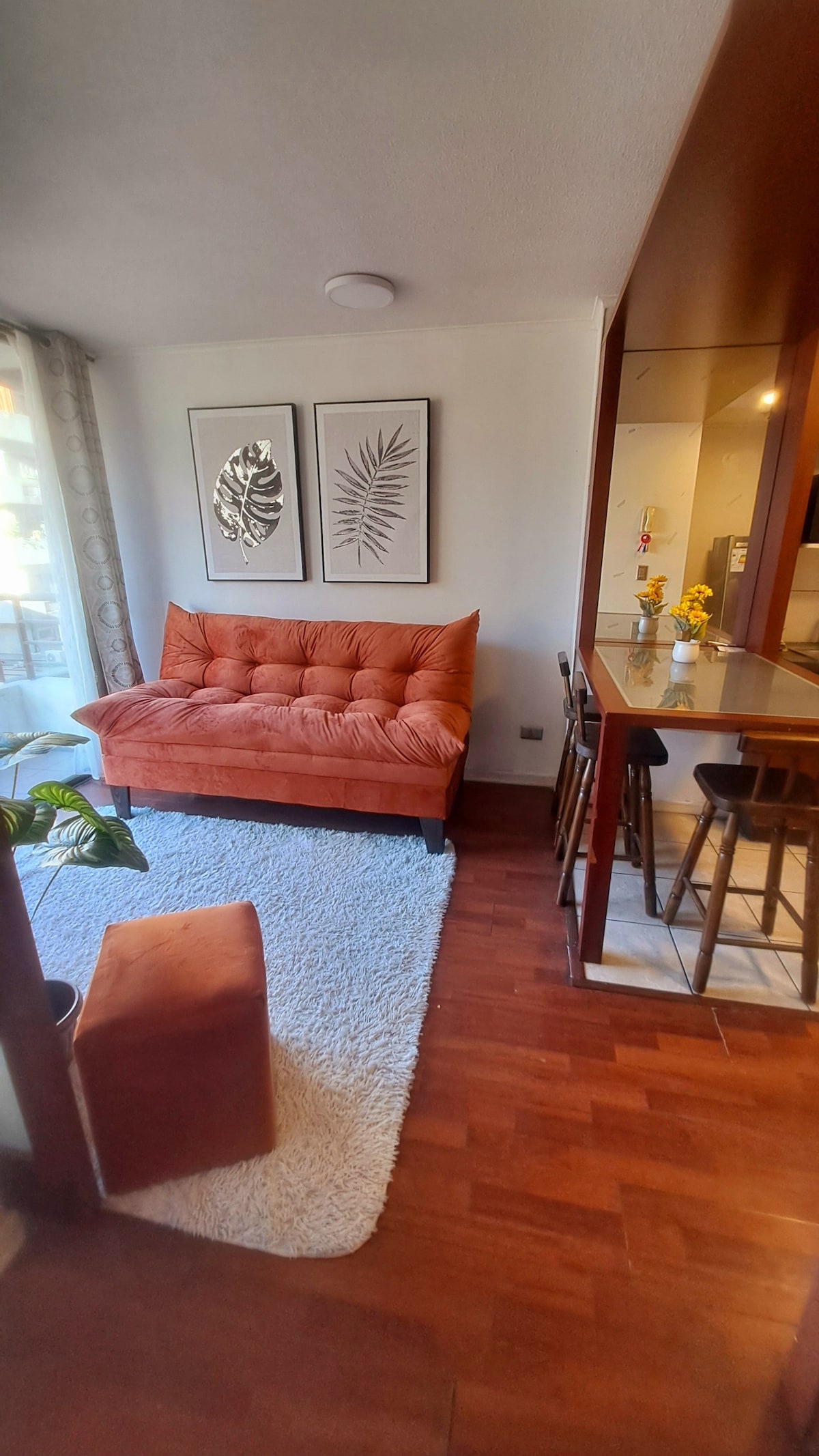 A warm-toned seating area is furnished with a stylish orange sofa and an accompanying ottoman, placed on a soft white rug. Two framed botanical prints adorn the wall, while natural light floods in through a large window, creating an inviting ambiance.