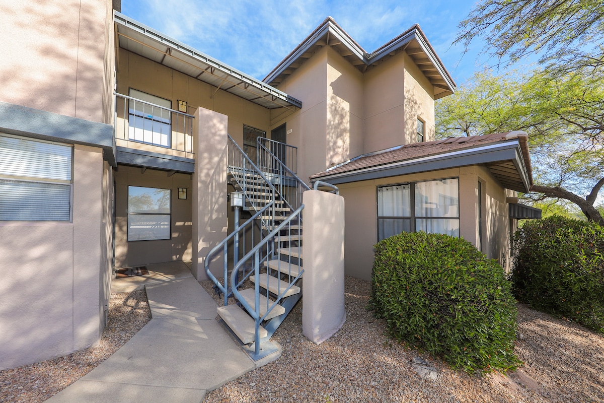 The exterior of the modern apartment building features a clean design with light-colored walls and a staircase leading to the upper levels. The landscape includes sparse gravel and shrubs, complementing the architecture under a clear blue sky.