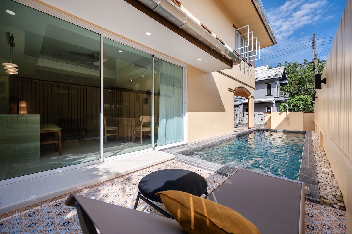 A serene outdoor space features a private swimming pool bordered by a sun terrace, equipped with lounge chairs. Large glass doors provide a view into the well-lit interior, enhancing the open layout. Natural light fills the area, complemented by clear blue skies overhead.