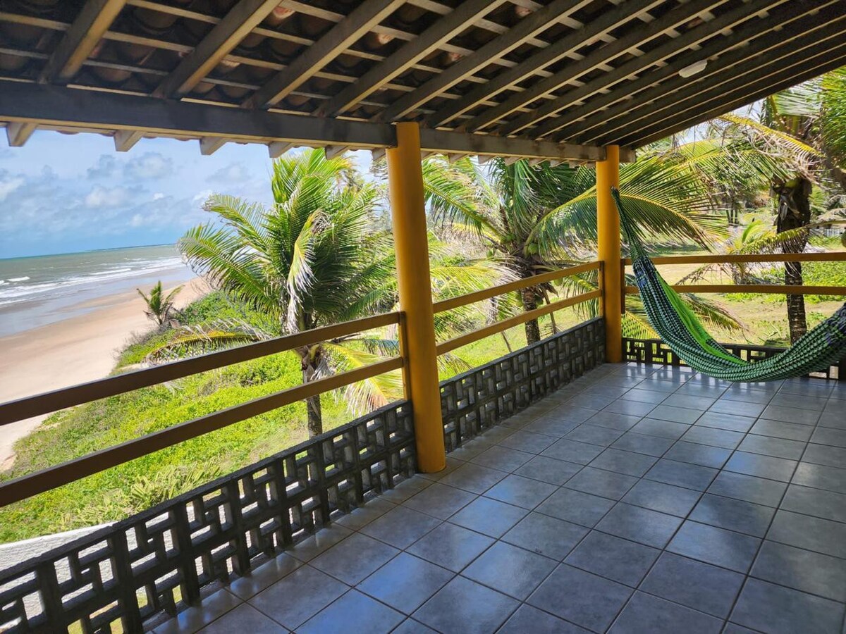 A spacious outdoor balcony is framed by wooden beams and features a hammock for relaxation. Lush palm trees are visible on either side, with a serene view of the sandy beach and ocean in the distance. Bright sunlight filters through the open space.