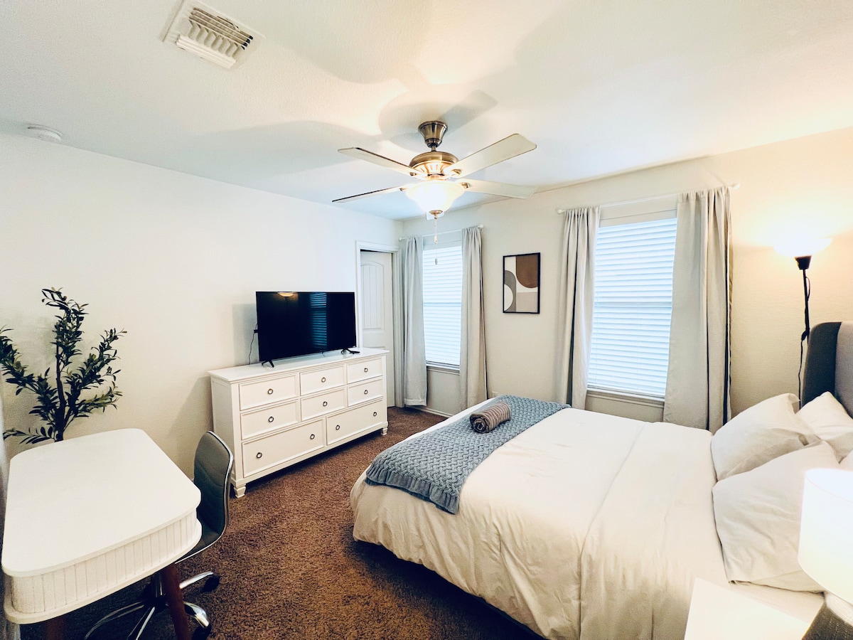 A modern private room is presented with a queen bed and a ceiling fan. A dresser is situated against one wall, accompanied by a wall-mounted TV. Two windows allow natural light to illuminate the space, while a work desk is positioned near the door.