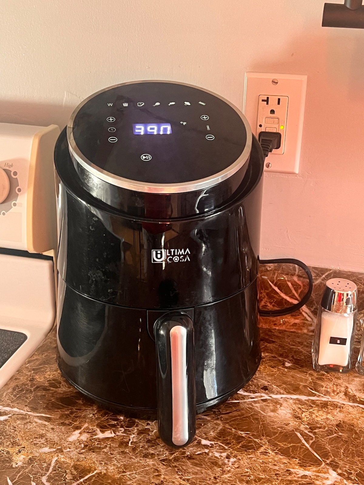 An air fryer is positioned on a marbled countertop, featuring a digital display panel with illuminated controls. Nearby, a salt shaker adds a functional element to the kitchen space. The appliance is sleek and modern, complementing the overall decor.
