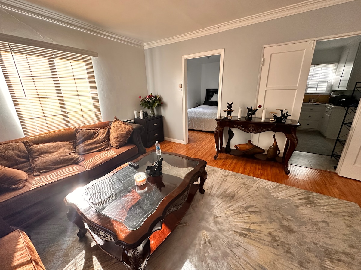 A cozy living area features a plush sofa and an ornate coffee table. Natural light filters in through a window with horizontal blinds, illuminating the room. A dining area can be seen through an open door, leading to a modern kitchen with contrasting cabinetry.