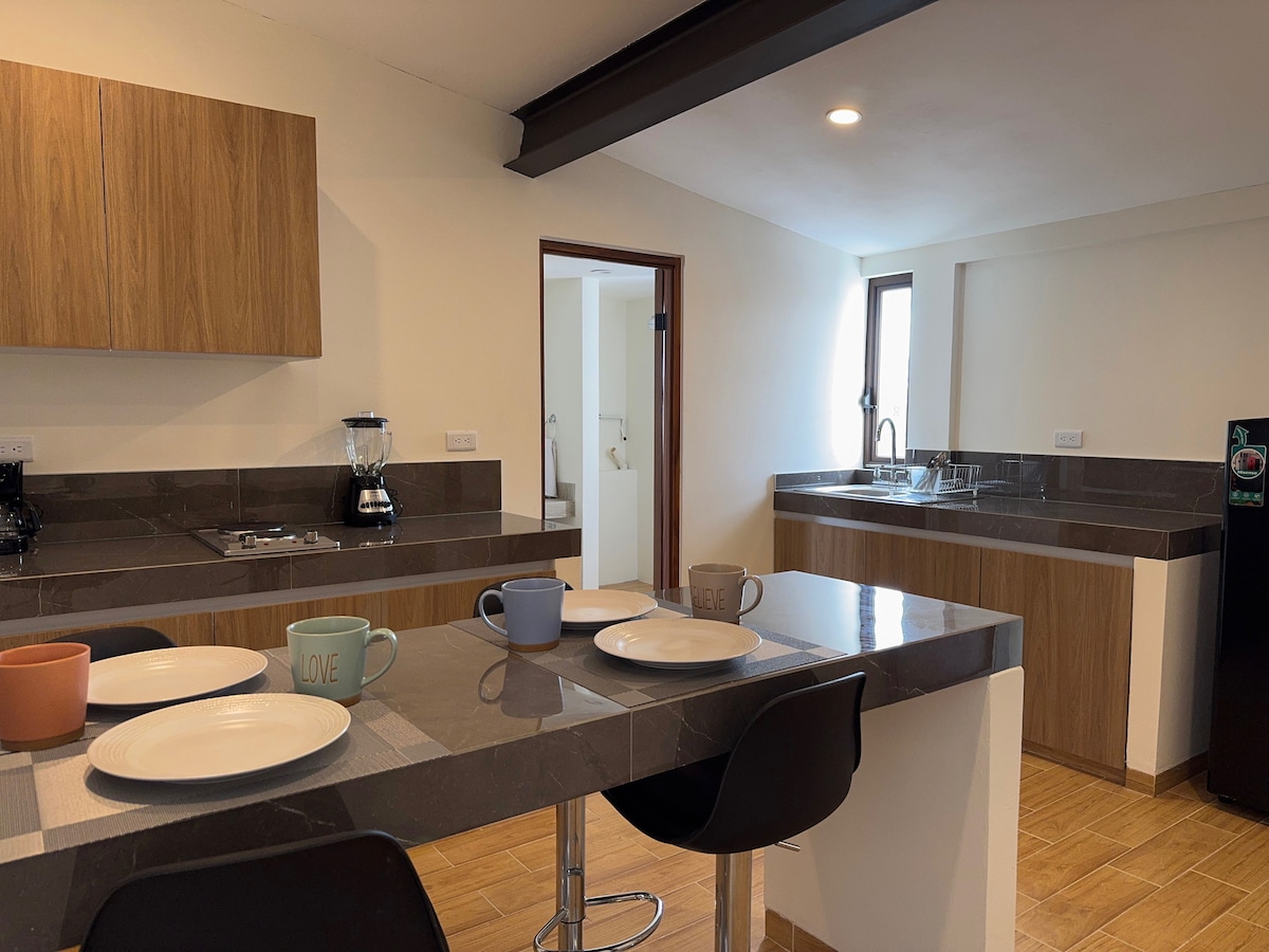 A modern kitchen space features wooden cabinetry and a sleek countertop. A coffee maker and electric kettle are visible, alongside plates and mugs set on the table. Natural light filters in through a window, creating a bright and functional cooking area.
