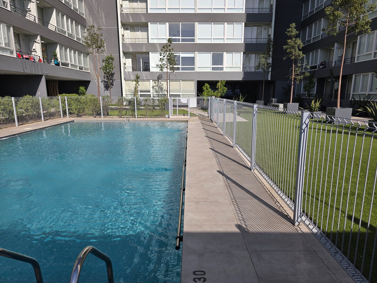 A swimming pool is framed by a clean walkway and fenced area, surrounded by well-maintained greenery. The water's surface reflects light, while the building's modern architecture provides a neutral backdrop. Lounge space is available nearby for relaxation.