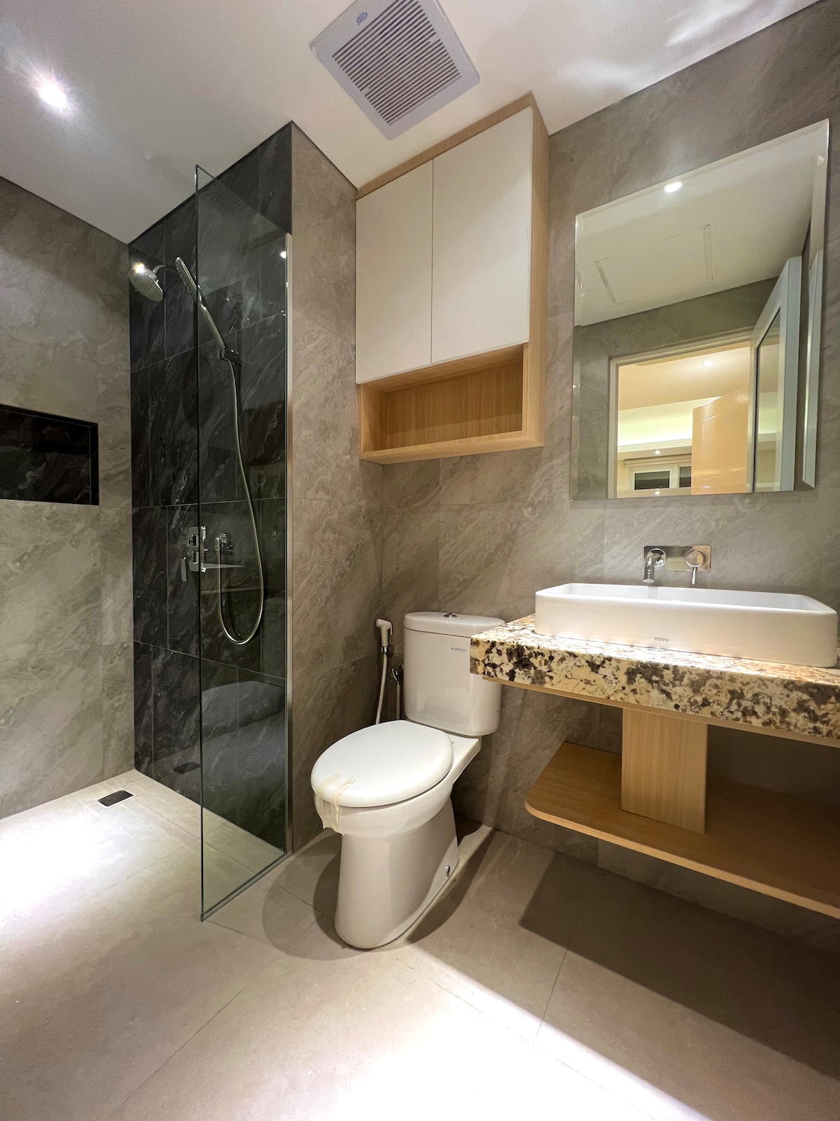 A modern bathroom features sleek marble walls and a glass-enclosed shower. A stylish vanity with a granite countertop is complemented by a simple mirror above. The space is well-lit, showcasing contemporary fixtures and an integrated storage solution.