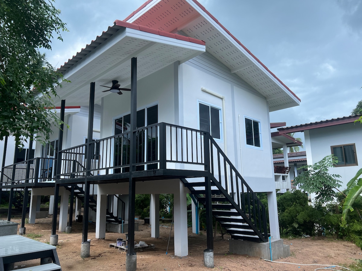 A two-story building is presented, elevated on pillars with a staircase leading to a covered porch. White walls and contrasting black railings create a modern appearance. Surrounding greenery and gravel paths suggest a serene, natural setting.
