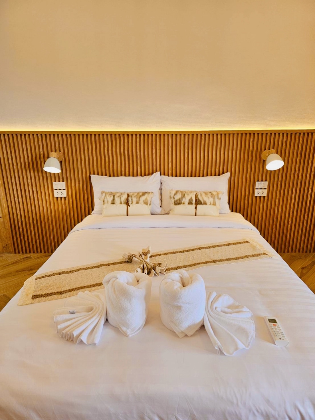 A spacious bed is centered in the room, adorned with soft pillows and a decorative blanket. Warm wooden panels create a cozy backdrop, while light fixtures on either side add a gentle glow. Two neatly arranged towels are folded at the foot of the bed.