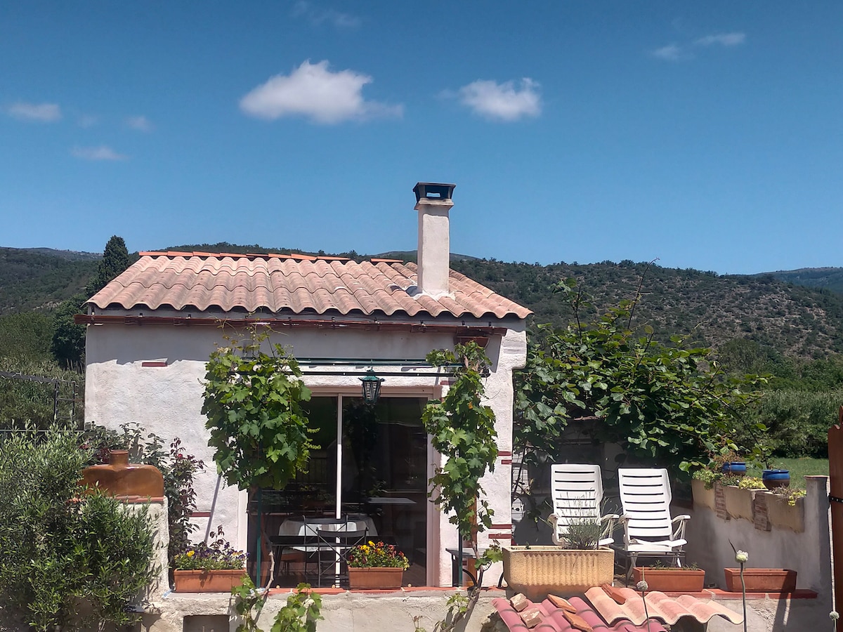 A charming white cottage features a terracotta roof and large glass doors, opening to a scenic outdoor area. Flanking the entrance are potted plants and a small table with chairs, while reclining chairs are positioned nearby, providing a tranquil view of the surrounding hills.