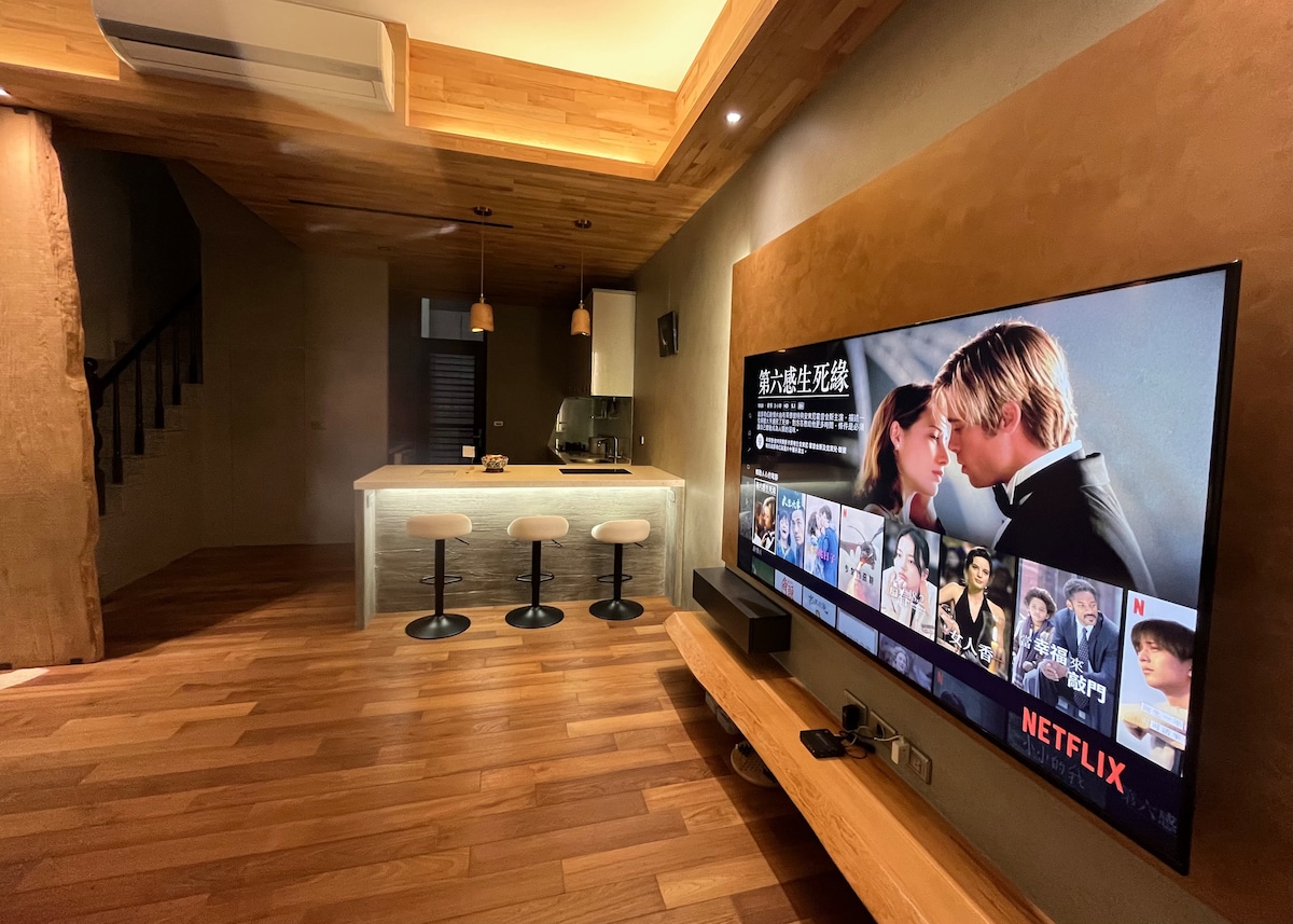 An open living space features a large flat-screen television mounted on the wall. A kitchen island with three bar stools is visible, complemented by wood accents and warm lighting. The floor has a polished wooden finish, enhancing the modern aesthetic of the area.