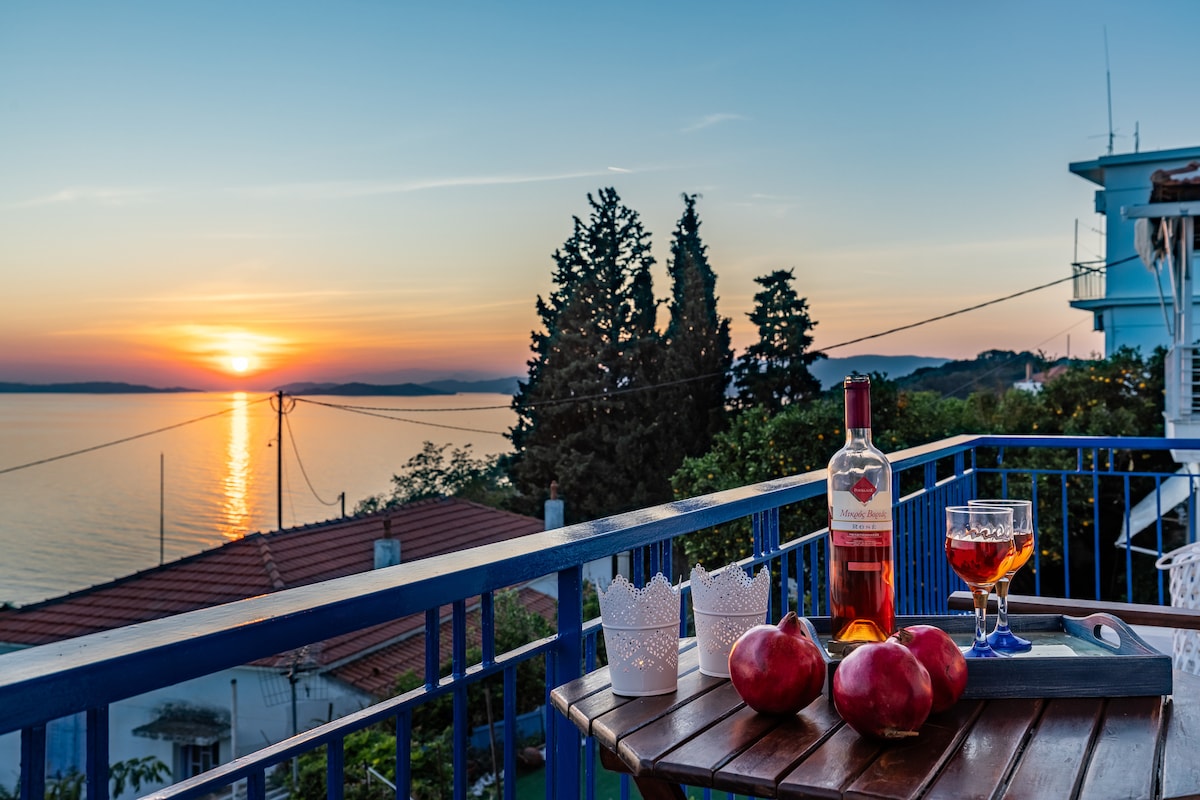 A serene balcony setting is captured at sunset, featuring a wooden table adorned with apples and a bottle of wine. The distant horizon is painted in warm hues as the sun sets over the water, illuminating nearby trees and rooftops.