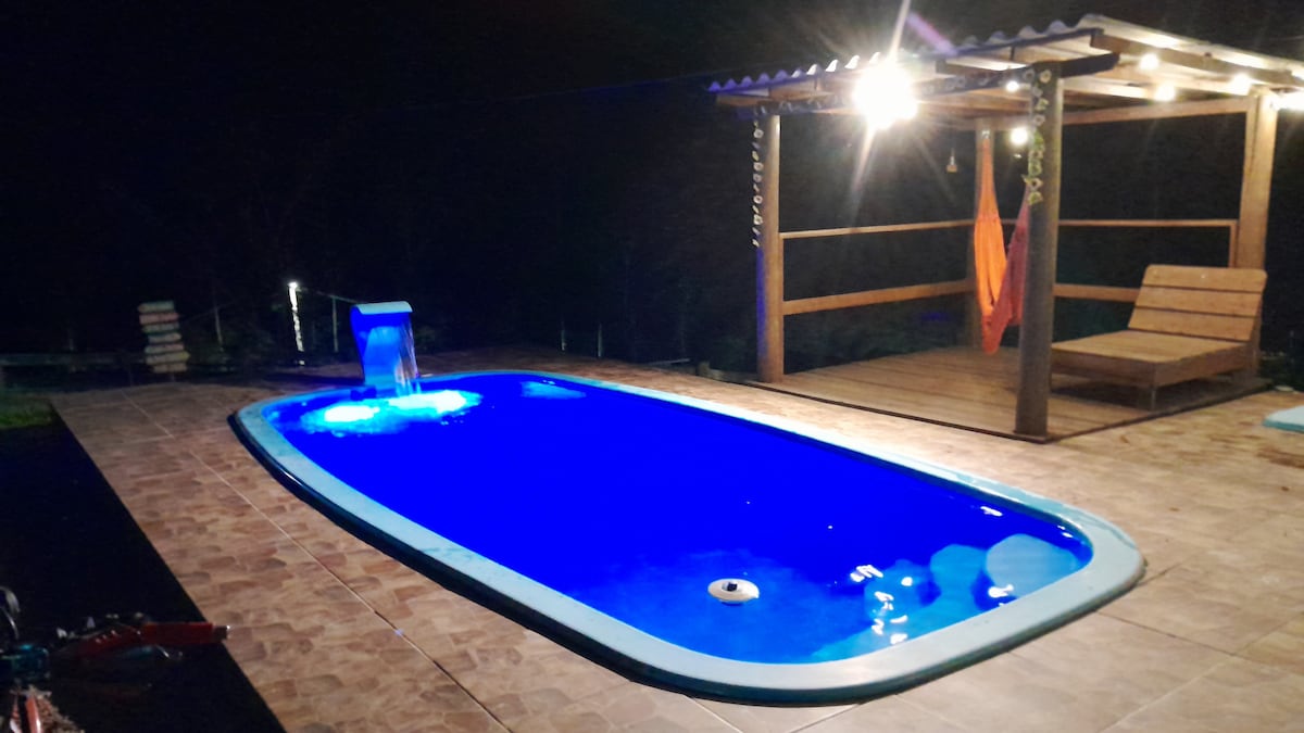 A rectangular pool features illuminated blue water, complemented by a gentle waterfall and a wooden deck. A hammock and a lounge chair are positioned nearby, surrounded by a tranquil outdoor space that is enhanced by soft lighting.
