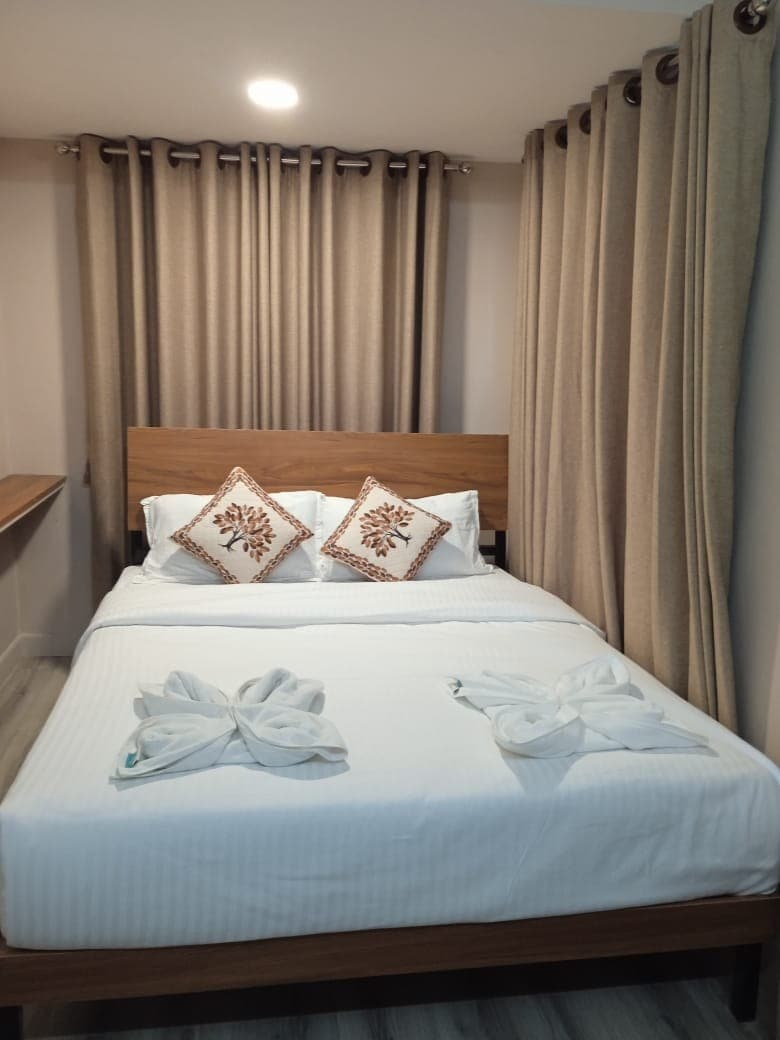 A well-lit bedroom features a spacious bed adorned with neatly arranged towels. Two decorative pillows with leaf patterns are placed at the back of the bed. Soft beige curtains frame the window, complementing the light colors of the room's decor.