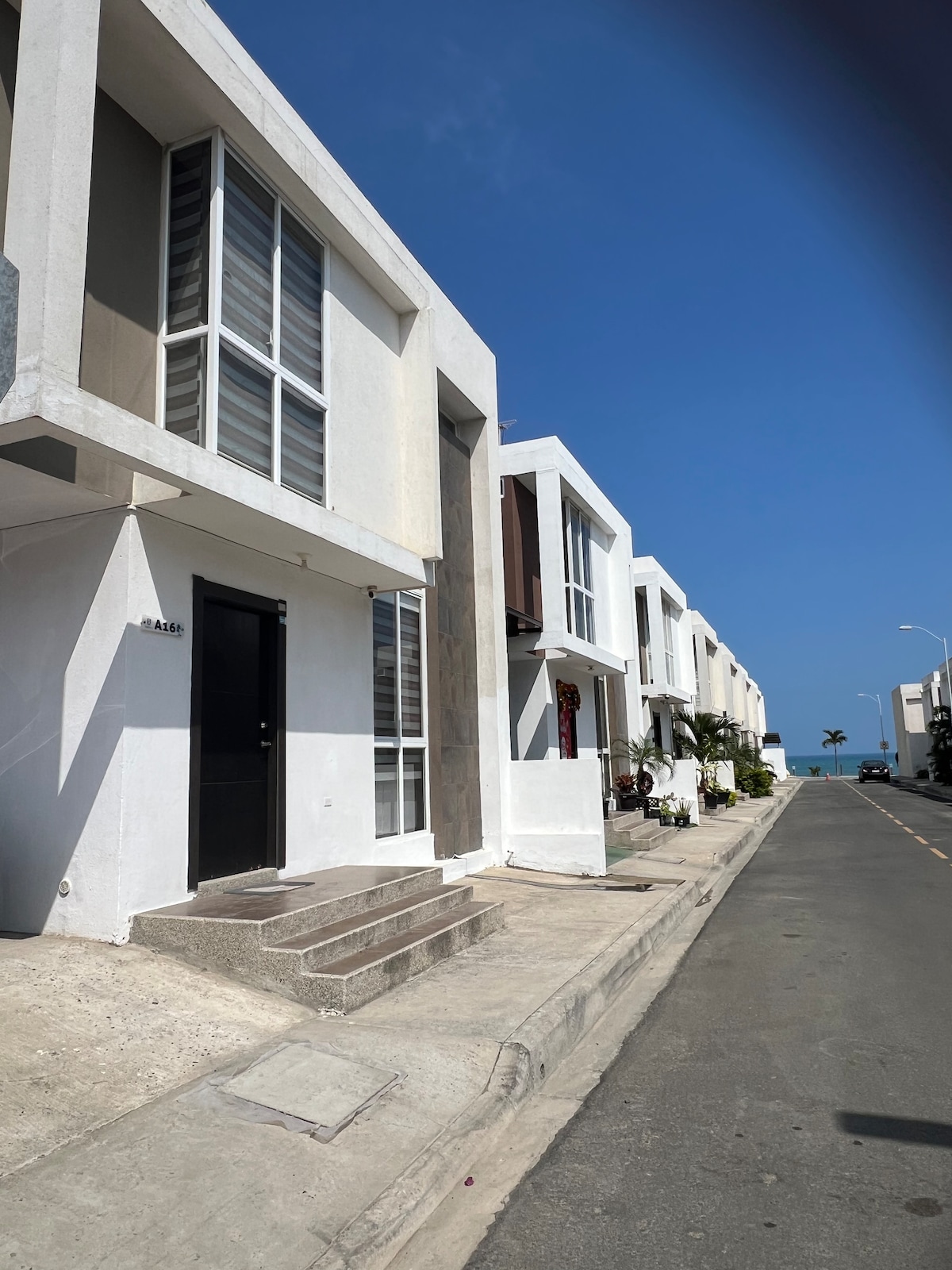 A quiet street is lined with modern houses featuring clean lines and large windows, allowing natural light to fill the interiors. Steps lead to an entrance that may be accessed easily. A glimpse of the ocean is visible at the end of the road, enhancing the serene setting.