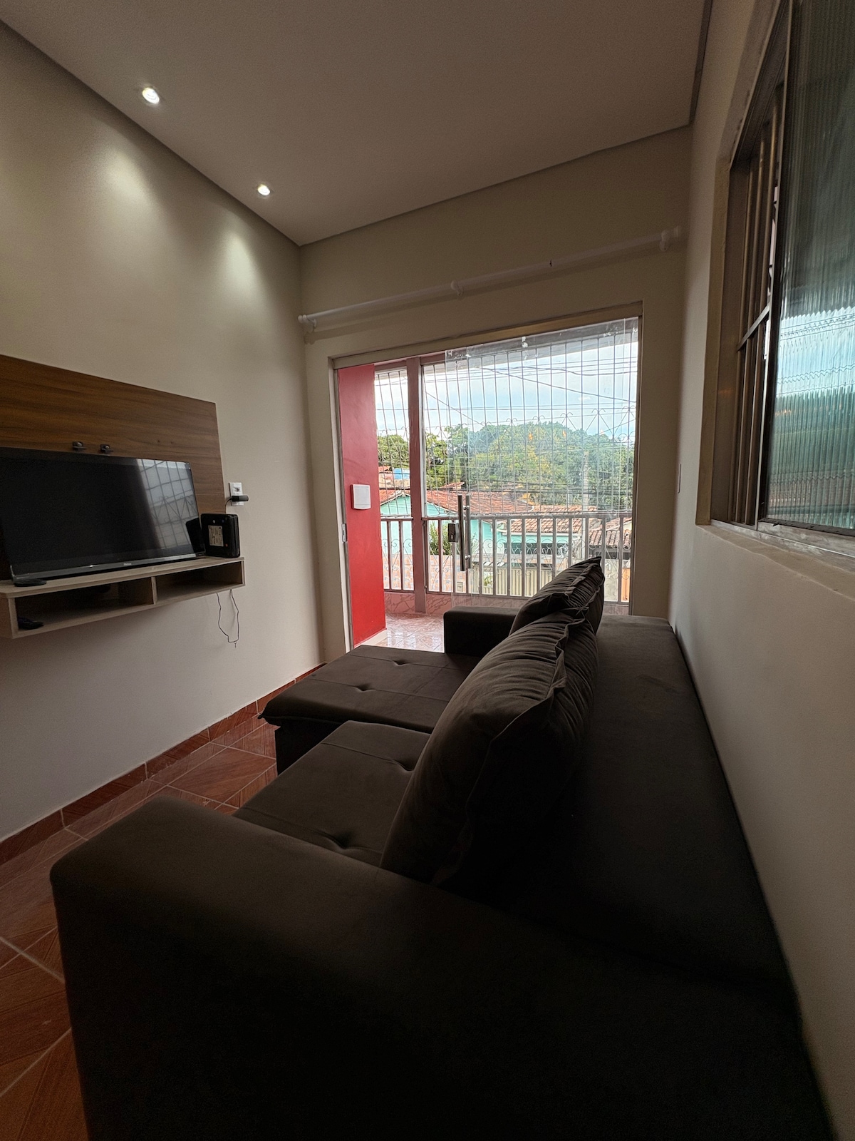 A modest living area features a dark sofa positioned against a wall with a mounted television. A small coffee station is available on a shelf. Natural light enters through a door leading to a balcony, which overlooks a scenic view of mountains and greenery.