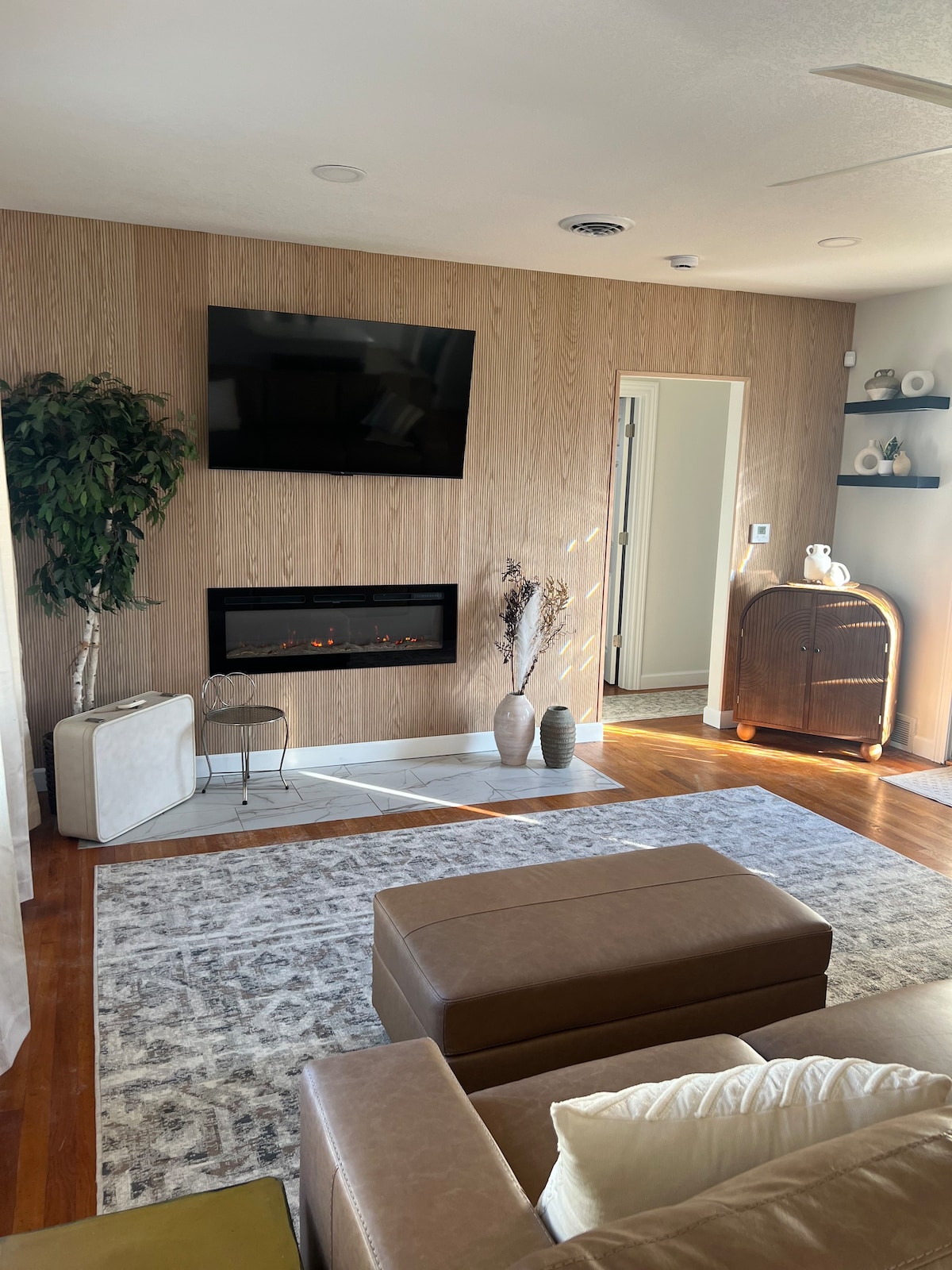 A cozy living area features a modern electric fireplace set against a wooden accent wall. A large area rug covers the wooden floor, while a flat-screen TV is mounted above the fireplace. Natural light enters through the nearby window, enhancing the serene atmosphere.