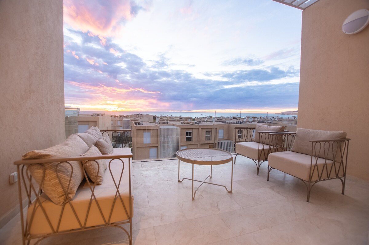 A spacious terrace features two comfortable seating arrangements facing a coffee table, all set against a vibrant sunset sky. The clean, minimalist design is enhanced by neutral tones and large glass panels, providing panoramic views of the surrounding area.