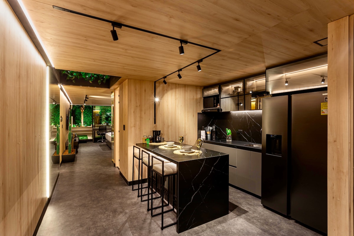 A modern kitchen space features sleek black countertops, illuminated by stylish under-cabinet lighting. Bar stools sit at a minimalist island, complemented by warm wood paneling that runs along the walls. Natural greenery is visible through large windows, creating a refreshing ambiance.