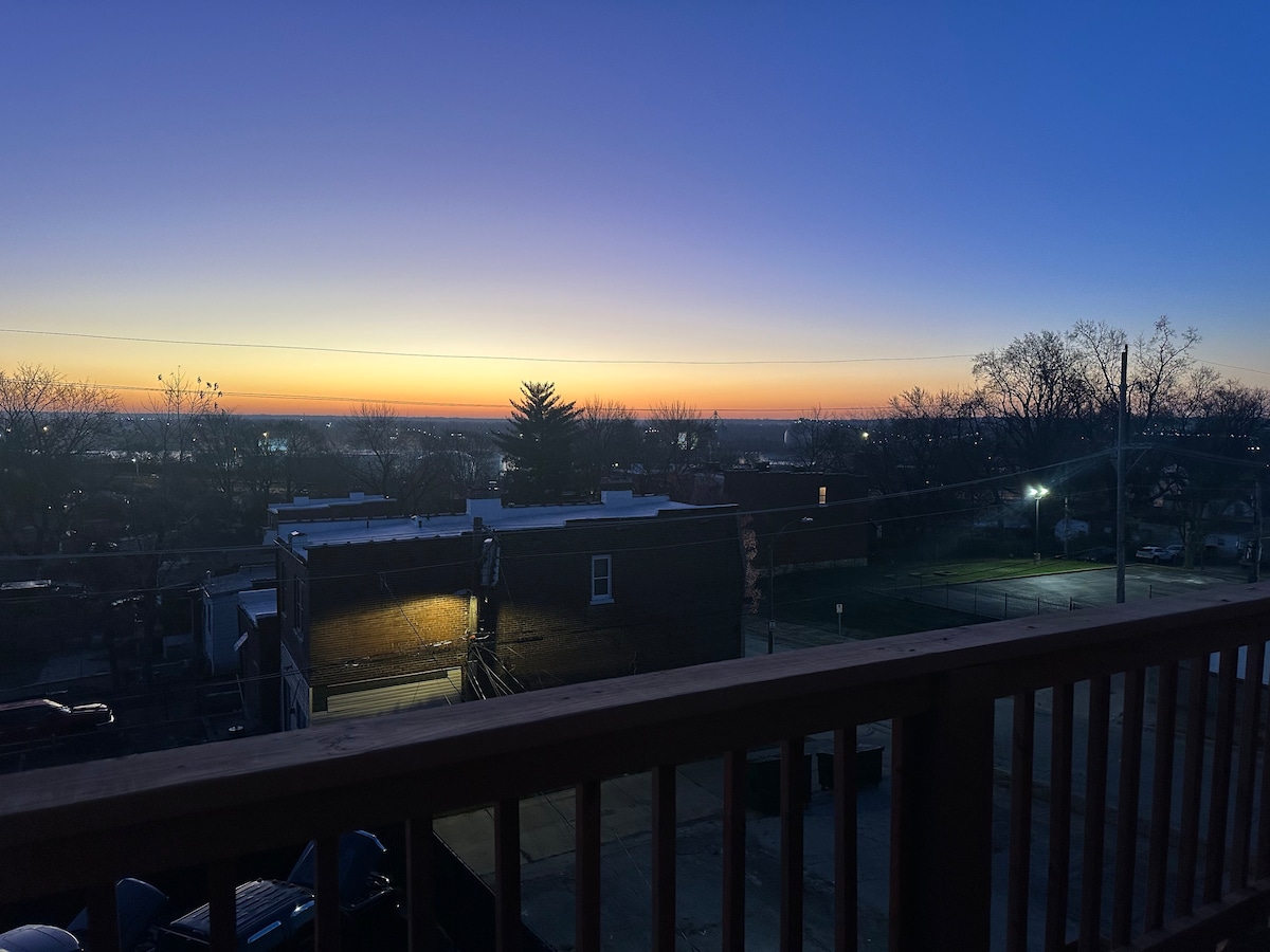 A serene view of a sunset skyline is captured from a balcony, showcasing a gradient of colors transitioning from deep blue to warm orange. The scene includes distant buildings silhouetted against the horizon, with soft lighting illuminating the nearby trees and parking area.