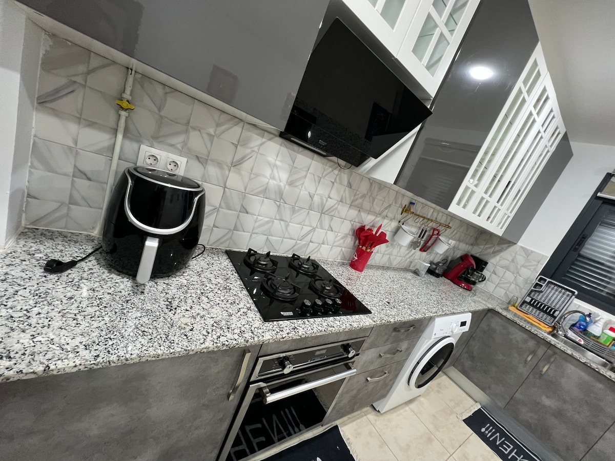 A modern kitchen area features sleek gray cabinets and a textured tile backsplash. Stainless steel appliances include a gas stove and an integrated oven. A black air fryer sits on the granite countertop, complemented by red utensils and a washing machine positioned near the end.