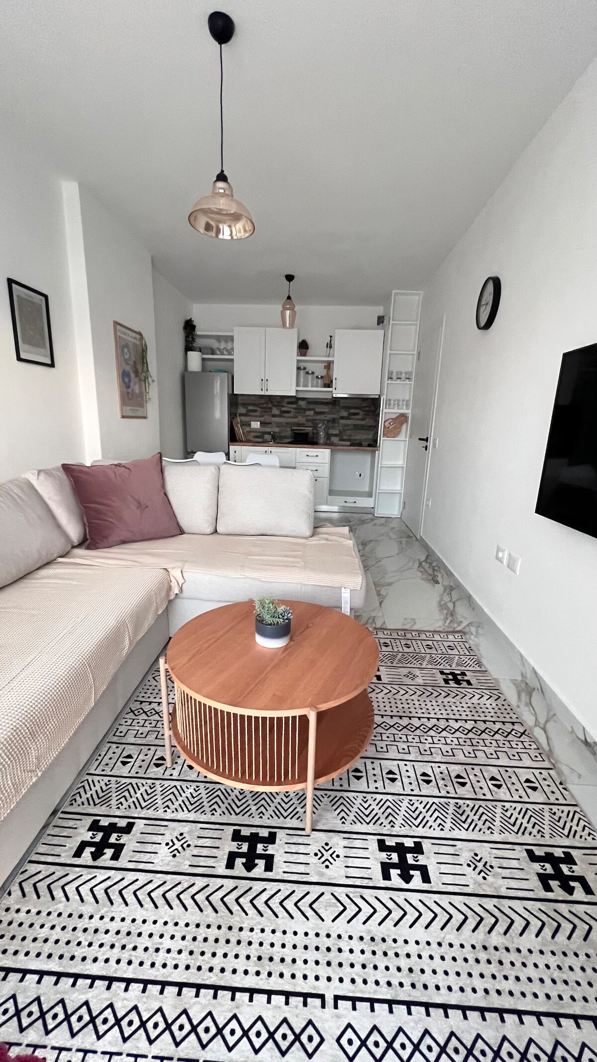 A bright living area features a corner sofa adorned with cushions in soft shades. A round wooden coffee table sits atop a patterned rug, with a small potted plant on display. The open kitchen area in the background showcases modern appliances and light cabinetry.