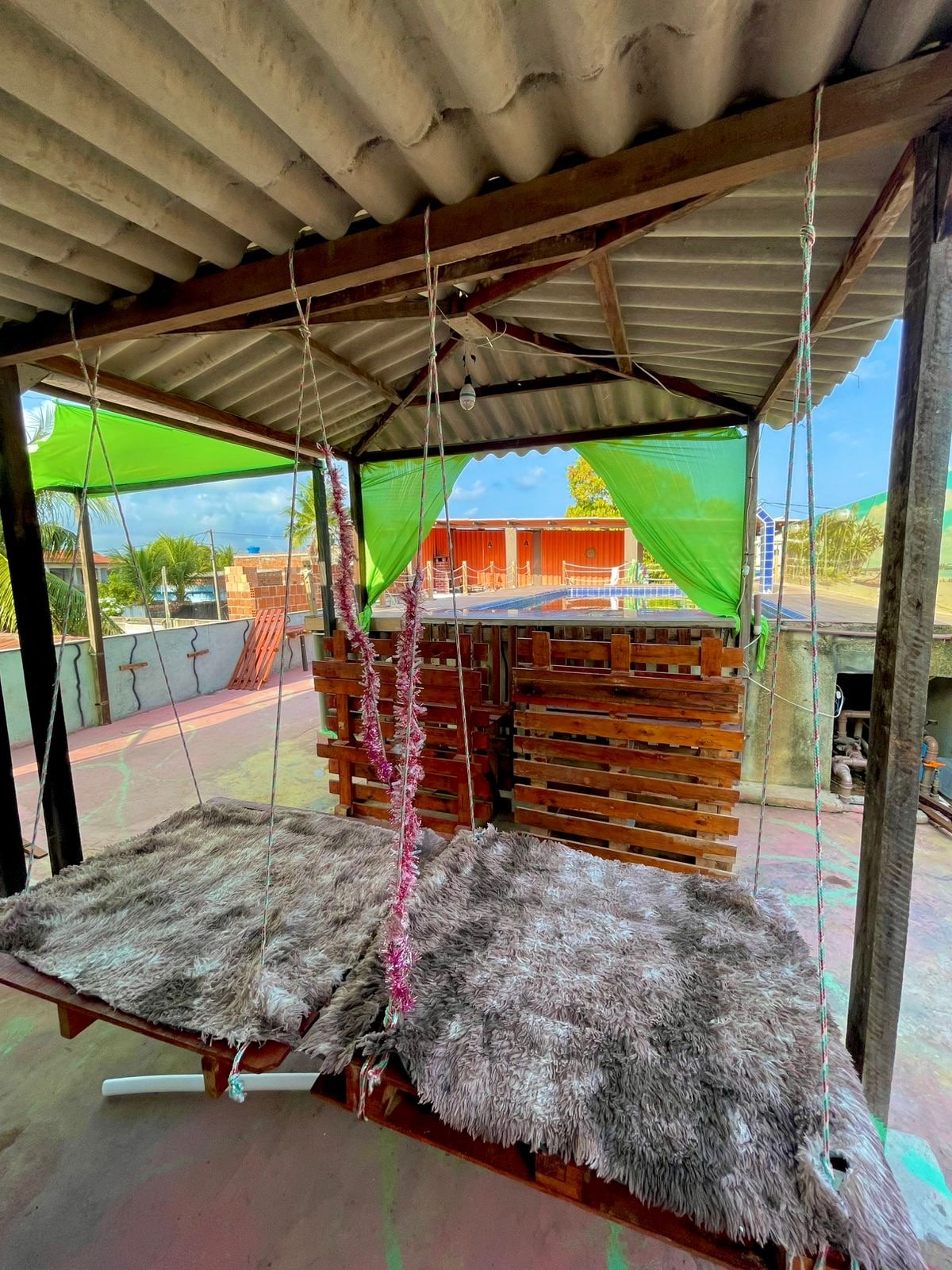 A shaded outdoor area features two swings adorned with soft, textured throws. Green fabric canopies provide shelter overhead, while wooden pallets create a rustic backdrop. The space invites relaxation and enjoyment of the surrounding atmosphere.