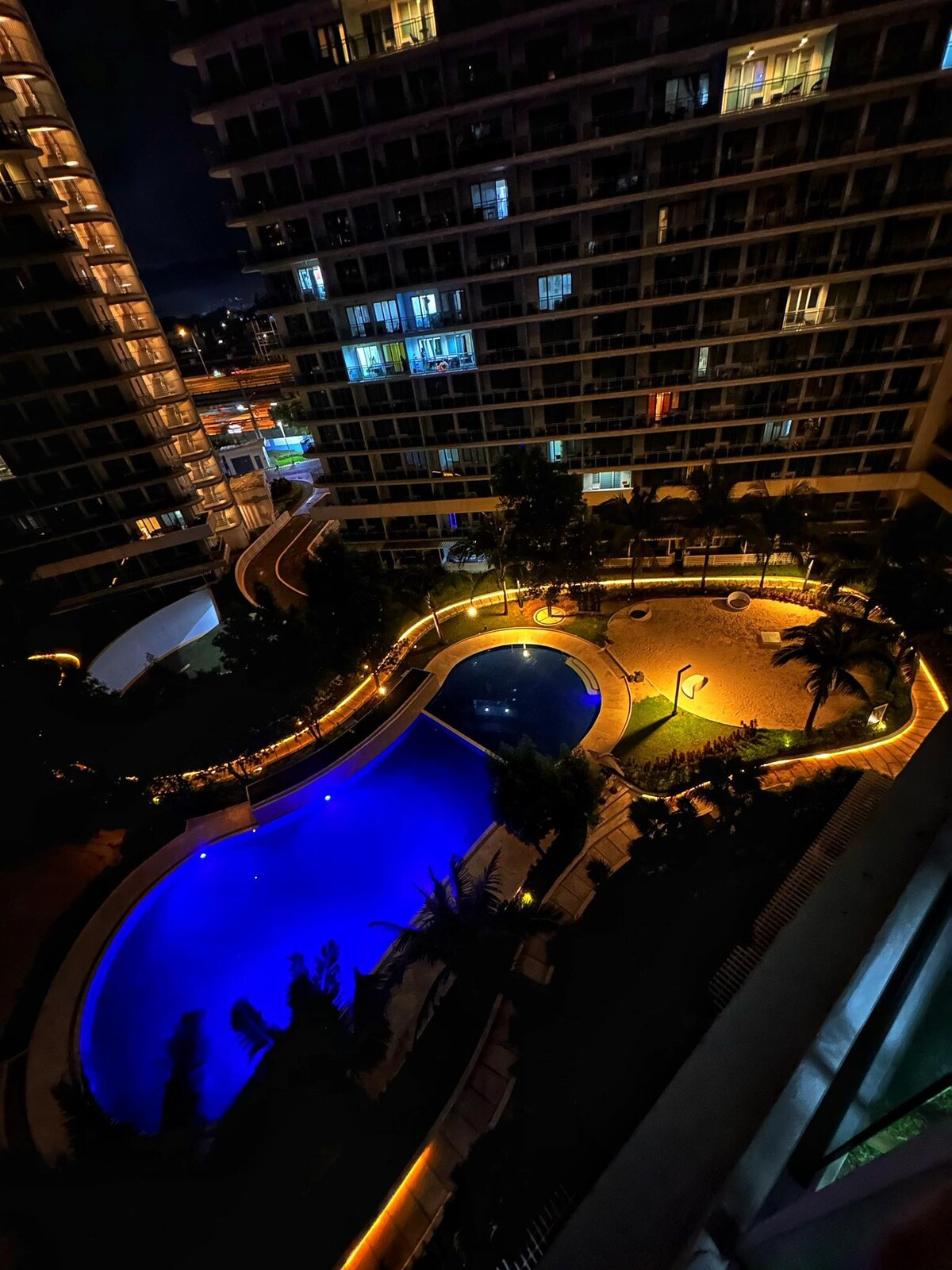 An aerial view captures the illuminated pool area at night, featuring two distinct pools surrounded by palm trees and landscaped gardens. Soft lighting outlines the pathways, enhancing the serene atmosphere of the outdoor space.