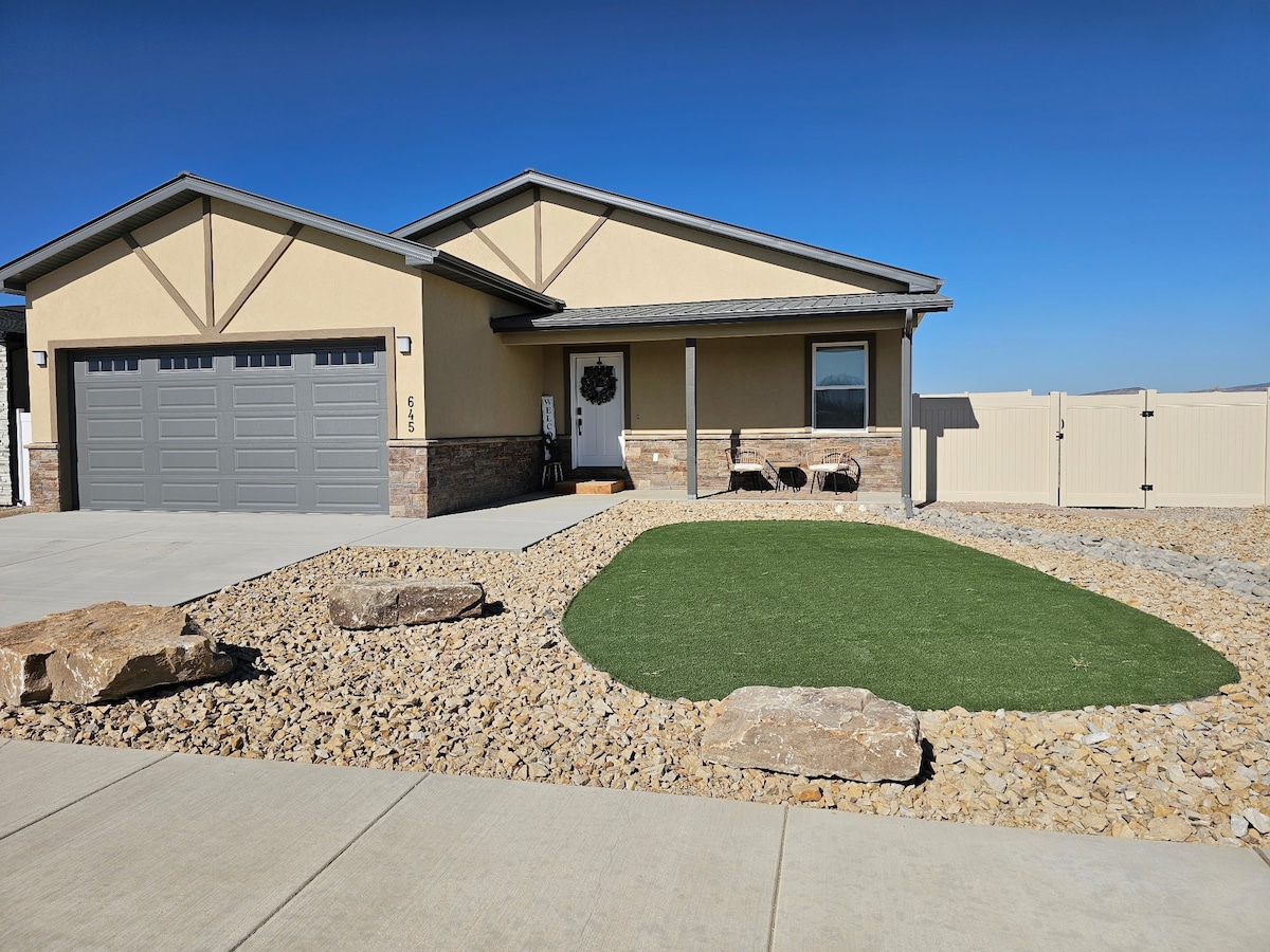A modern single-story home presents a smooth concrete driveway leading to a double garage. The tidy front yard features decorative stones and artificial grass, complemented by two chairs positioned near the entrance. A clear blue sky serves as a backdrop, enhancing the home's exterior.
