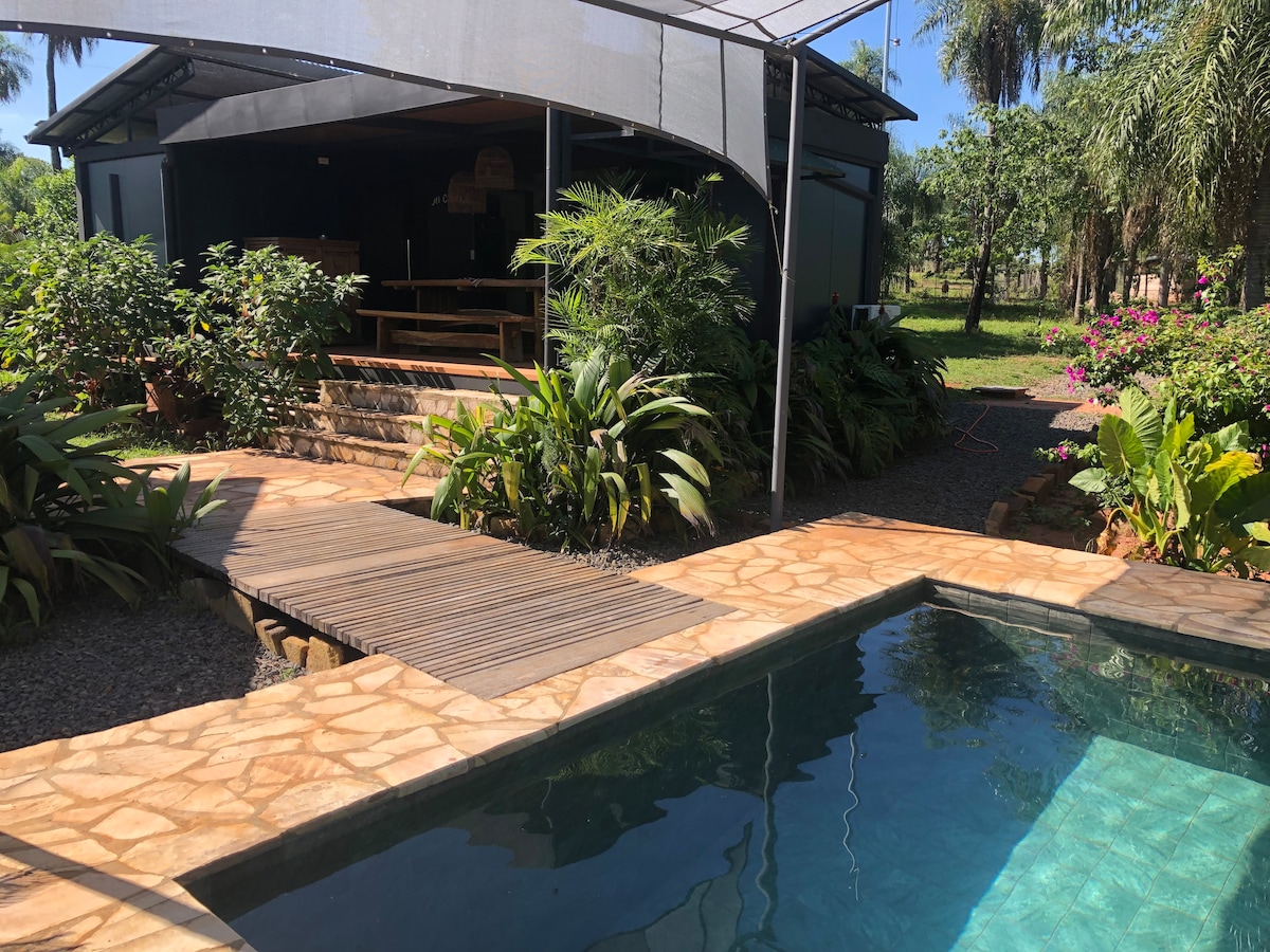 A stone pathway leads to a salt-water pool, surrounded by lush greenery and vibrant flowers. The pool reflects the clear sky, with a shaded area featuring wooden seating for relaxation. A cozy outdoor living space is visible in the background, enhancing the tranquil setting.