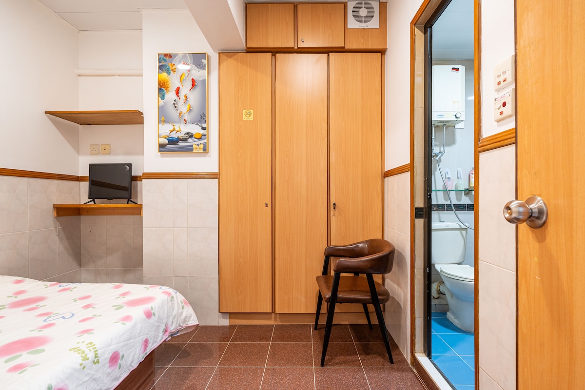 A compact room features a spacious bed with a floral patterned duvet. A wooden chair is positioned beside a small table with a television. Storage is provided by built-in wooden closets. A bathroom is visible through a glass door, showcasing tiled flooring and additional amenities.