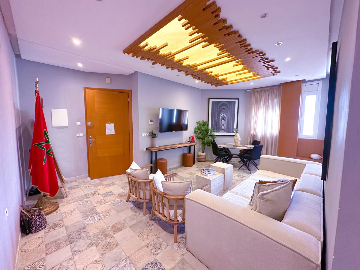 A modern living area features a neutral sofa facing a wall-mounted TV and an artistic wooden ceiling design. Natural light enters through large windows, illuminating a cozy seating arrangement with two chairs. A decorative plant adds a touch of greenery to the space.