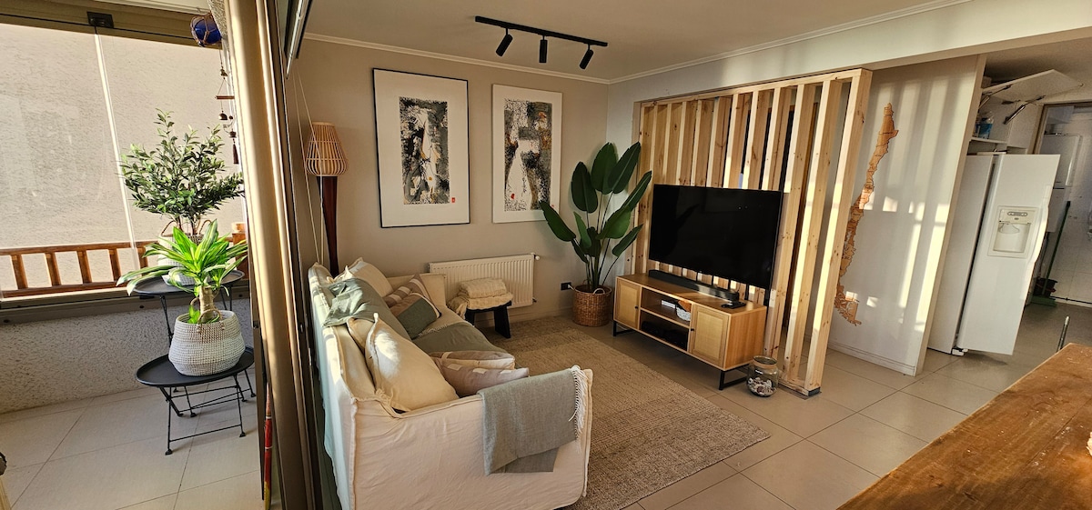 A living space is presented with a comfortable sofa and a wooden coffee table, complemented by decorative wall art. A large potted plant and a small tree contribute to the greenery. Natural light fills the area from a window, enhancing the inviting atmosphere.