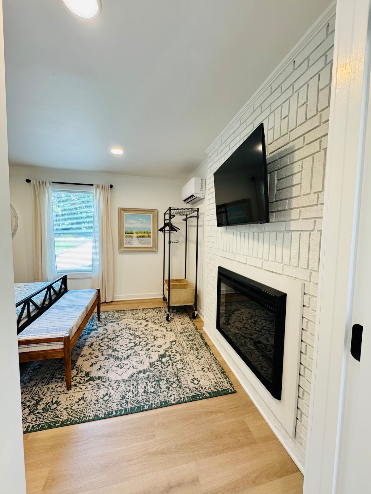A cozy entrance area features a spacious bed with a simple design beside a large window. A flat-screen TV is mounted on a textured white wall, while an electric fireplace adds warmth. An elegant area rug enhances the inviting ambiance of the space.