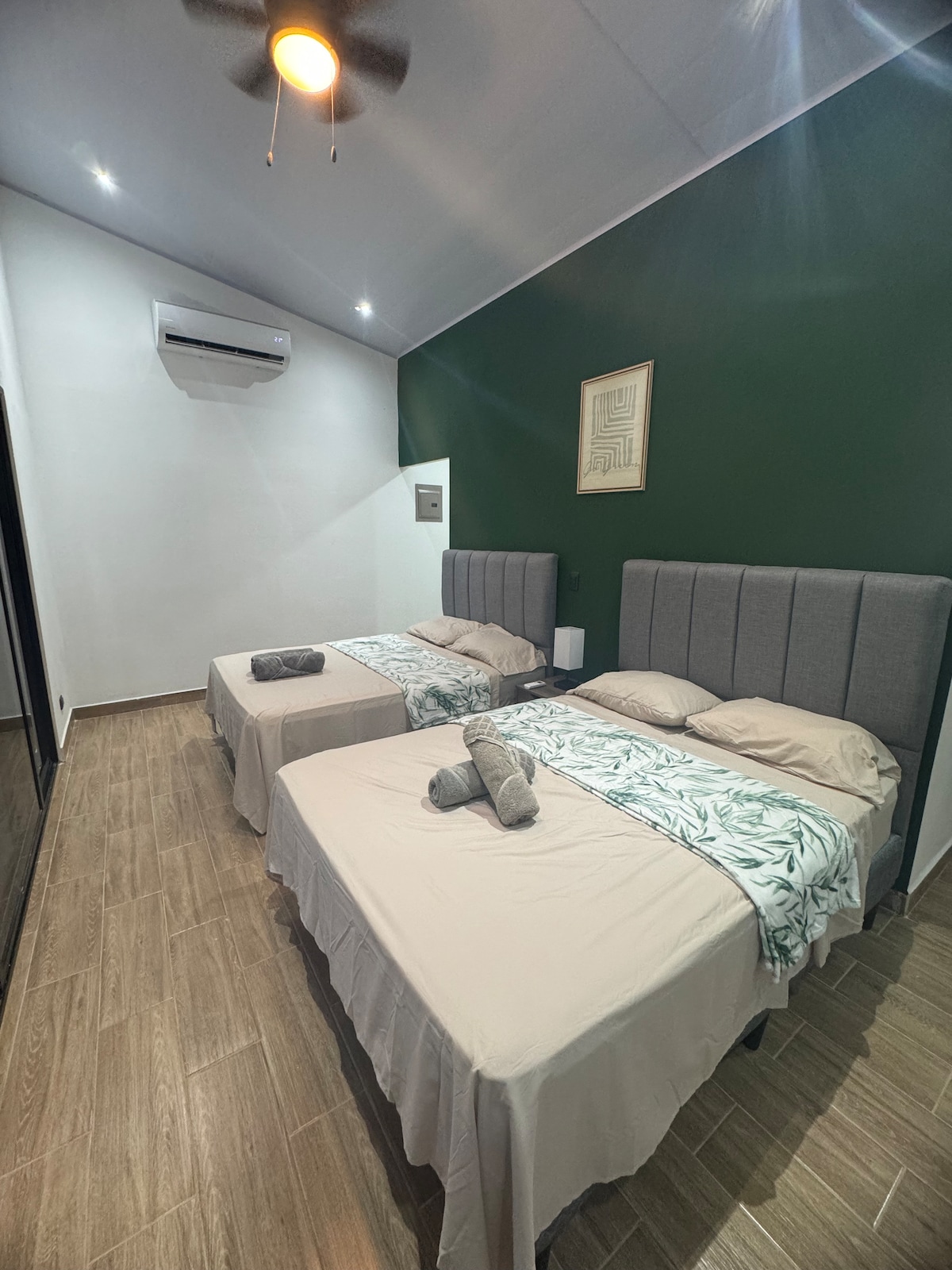 The room features two spacious beds adorned with neutral bedding and decorative towels. A rich green accent wall complements the light-colored flooring. Soft lighting enhances the cozy atmosphere, while a ceiling fan contributes to air circulation. An air conditioning unit is visible for added comfort.