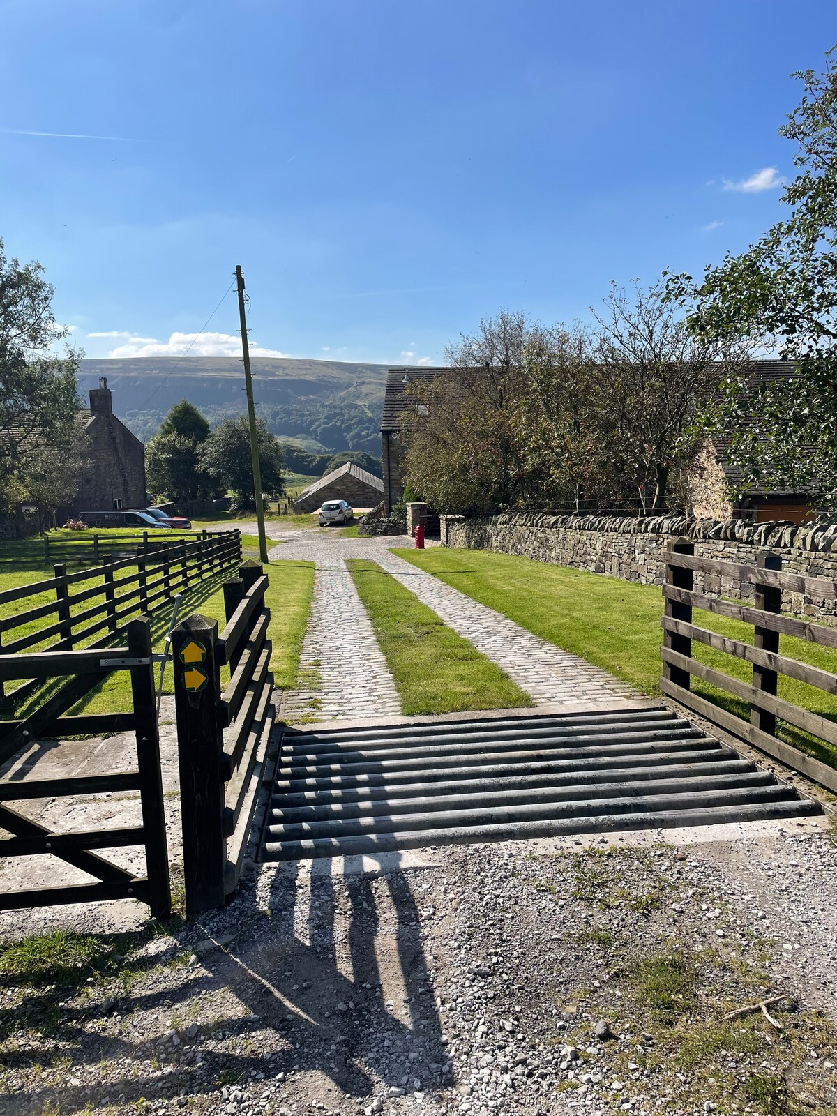 A gravel pathway leads towards a cottage, framed by grassy areas and bordered by stone walls. A wooden gate opens to the entrance, with distant hills visible under a clear blue sky. A few trees and a red postbox add to the rural charm of the surroundings.
