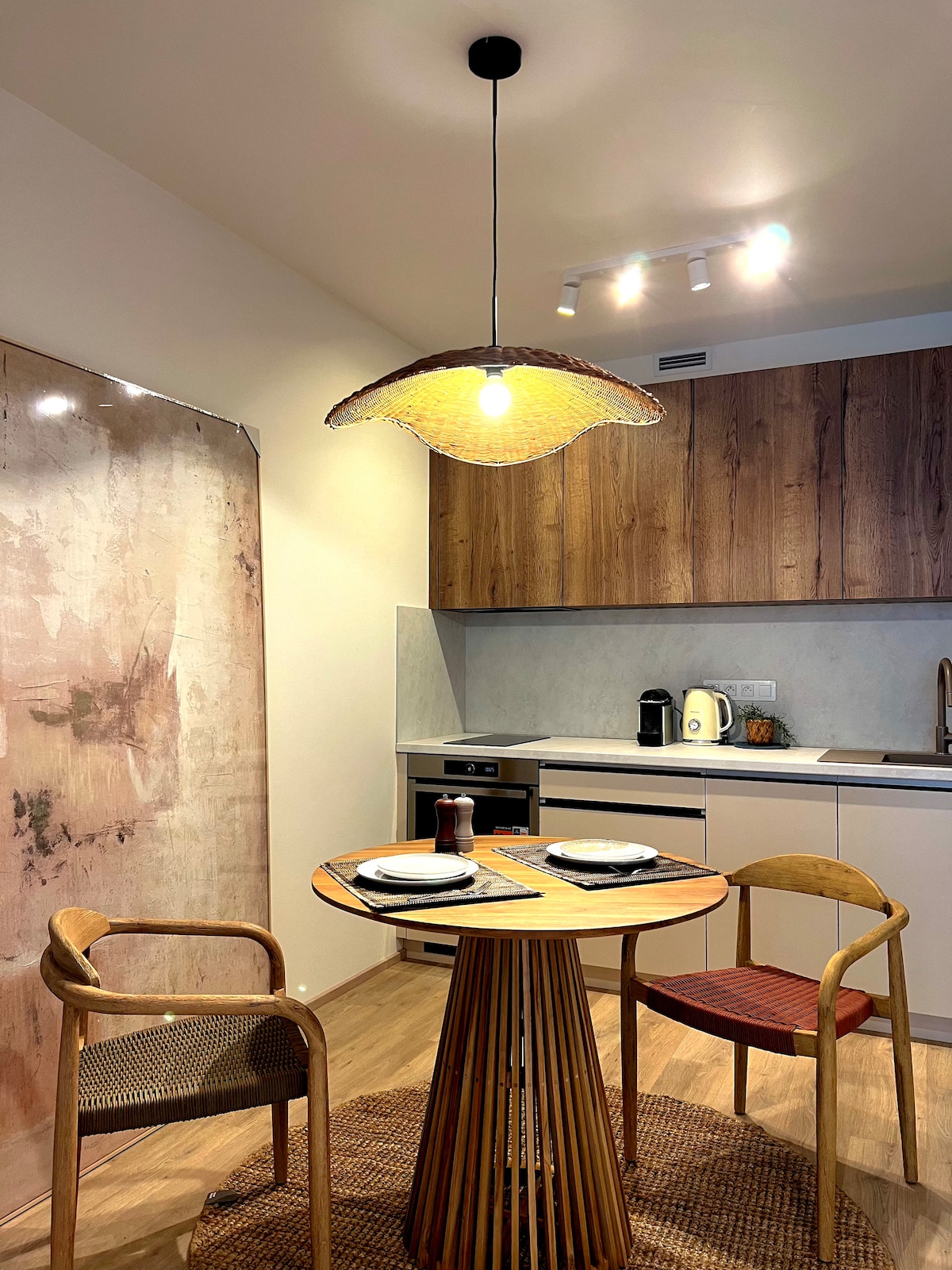 An inviting dining area is showcased, featuring a round wooden table set for two. Two chairs with different woven patterns are included, and a pendant light hangs above, providing warm illumination. The sleek kitchen cabinetry in a wood finish complements the modern design.