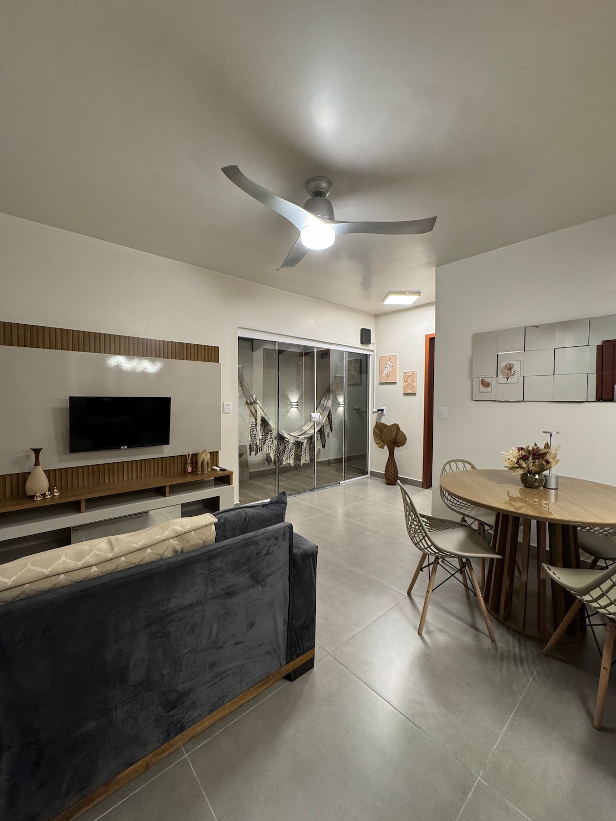 A living area features a comfortable sofa facing a wall-mounted television. A round dining table with modern chairs is positioned nearby, while sliding glass doors separate the space from a patio area. Light fixtures provide illumination, enhancing the room's clean and contemporary design.