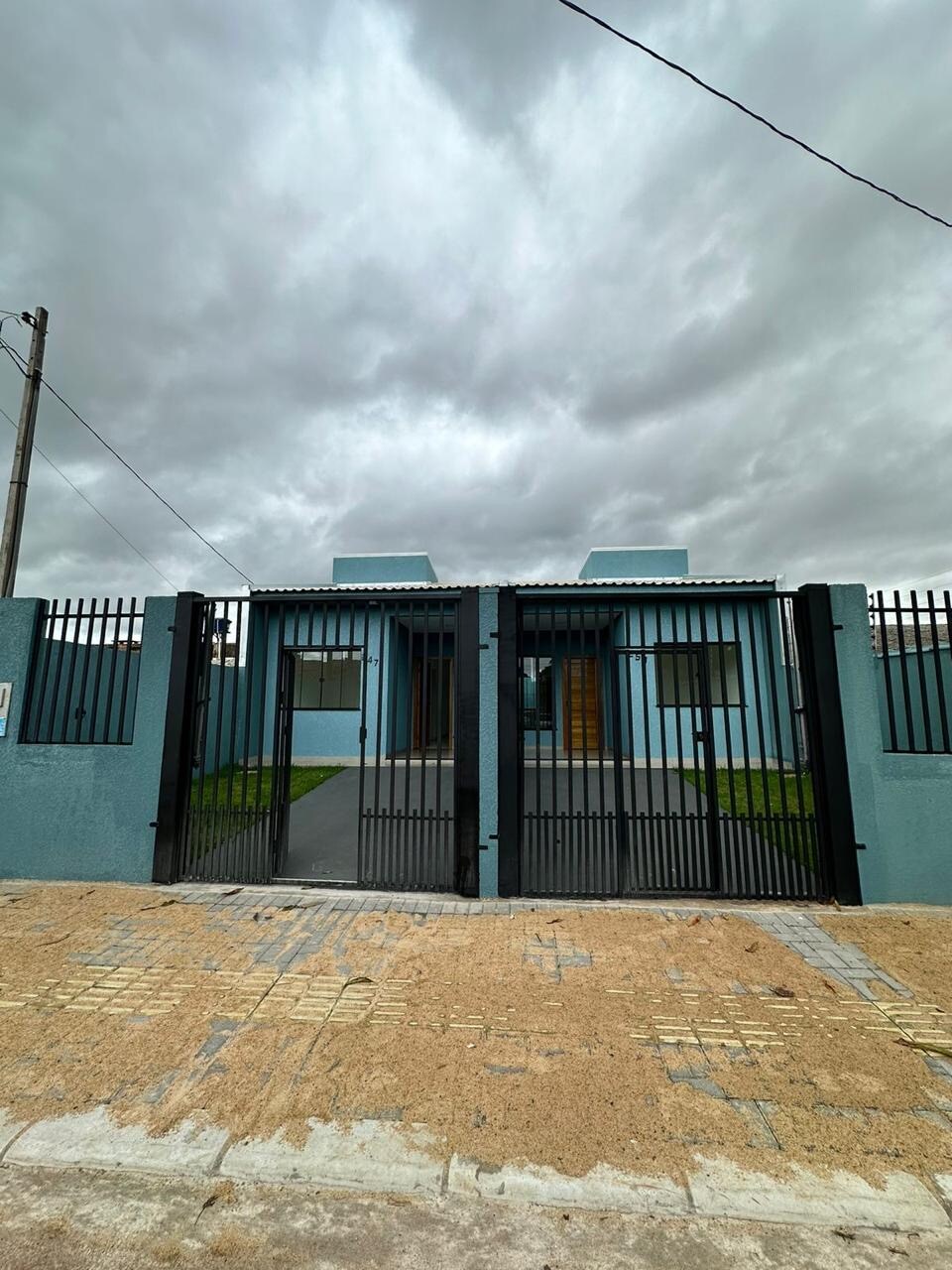 A modern facade is presented by the property, featuring a dual-gated entrance with black metal bars. The exterior is painted in soft blue tones, complemented by green landscaping. A paved pathway leads to the entrance, while the sky above displays a cloud-covered atmosphere.