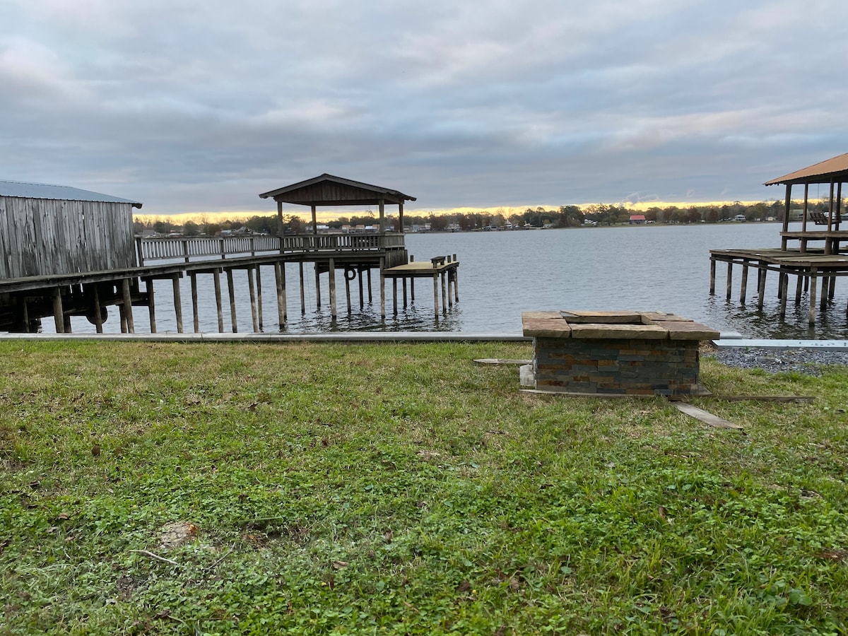 A serene waterfront view is shown, featuring two docks extending into the water. A fire pit made of stone is situated on the grassy shore, offering a place for gathering. The sky is overcast, with soft light reflecting on the water's surface.