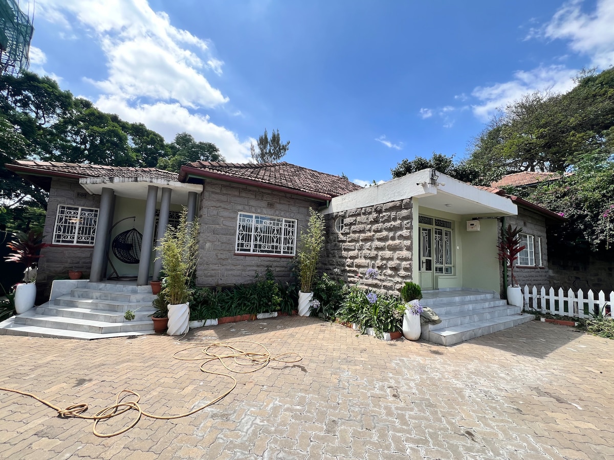 The entrance of a single-story bungalow is depicted, featuring a blend of stone and plaster walls. Steps lead to a covered porch with large glass doors. Lush greenery, including potted plants, enhances the welcoming appearance of the home, with a well-maintained lawn visible in the foreground.
