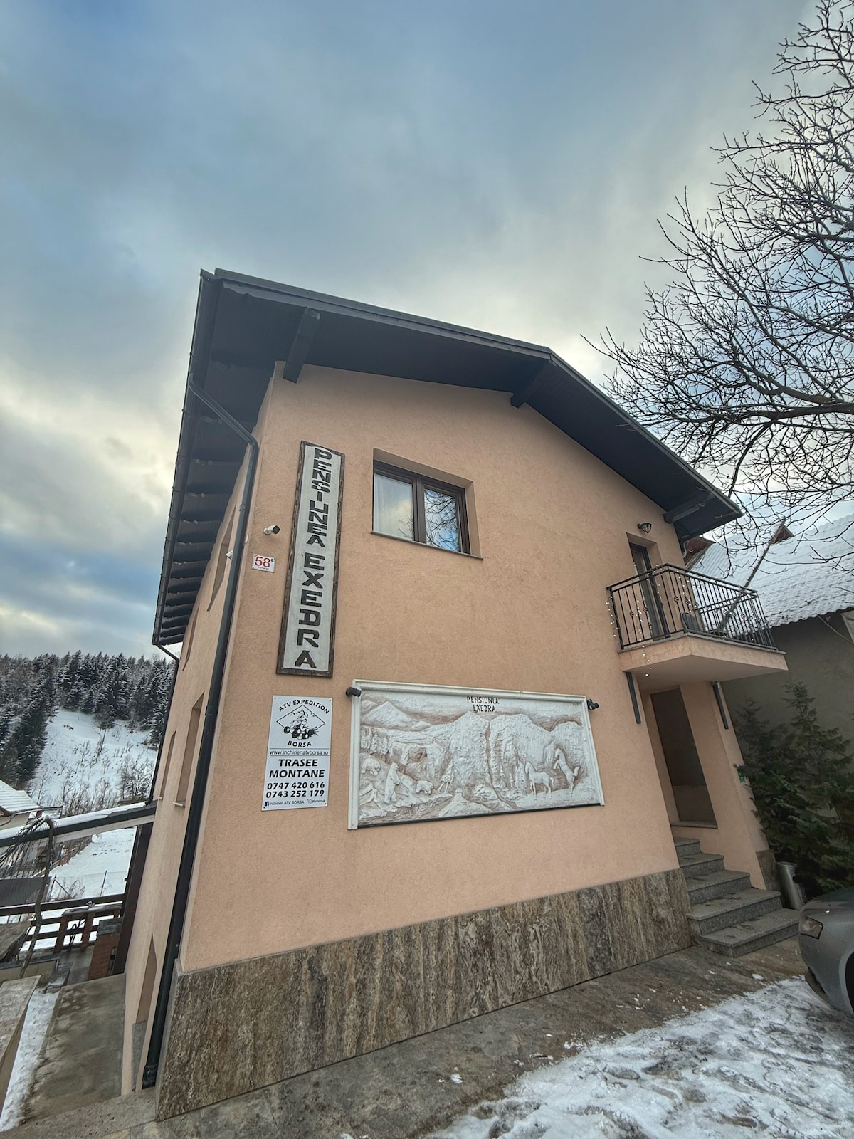 A two-story building with a light pink façade and stone accents is shown. A decorative wall mural is featured prominently, while a small balcony is positioned above the entrance. The surrounding area includes snow-covered ground and trees in the background.