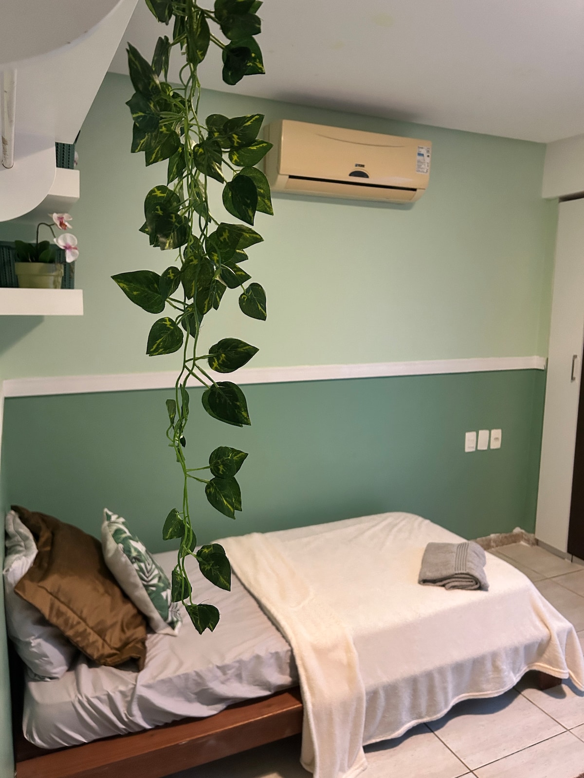 A cozy sleeping area features a spacious bed dressed in neutral linens, with a light blanket carefully placed. The serene green walls are complemented by a decorative shelf and an air conditioning unit, while a trailing plant adds a touch of nature.