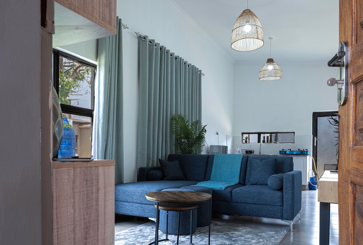A cozy living area features a blue sectional sofa with a soft throw draped over it. Natural light enters through the large window, complemented by sheer curtains. A round coffee table is centered on a patterned area rug, creating a welcoming space for relaxation.