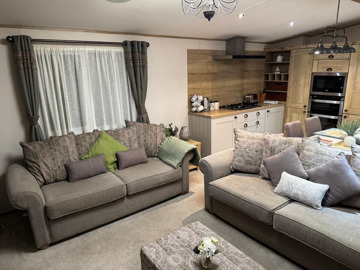 A cozy living area features two spacious sofas adorned with decorative cushions. A neutral-toned rug anchors the seating arrangement, while a light fixture hangs above. A well-equipped kitchen is visible in the background, with wooden cabinetry and counter space complementing the overall inviting atmosphere.