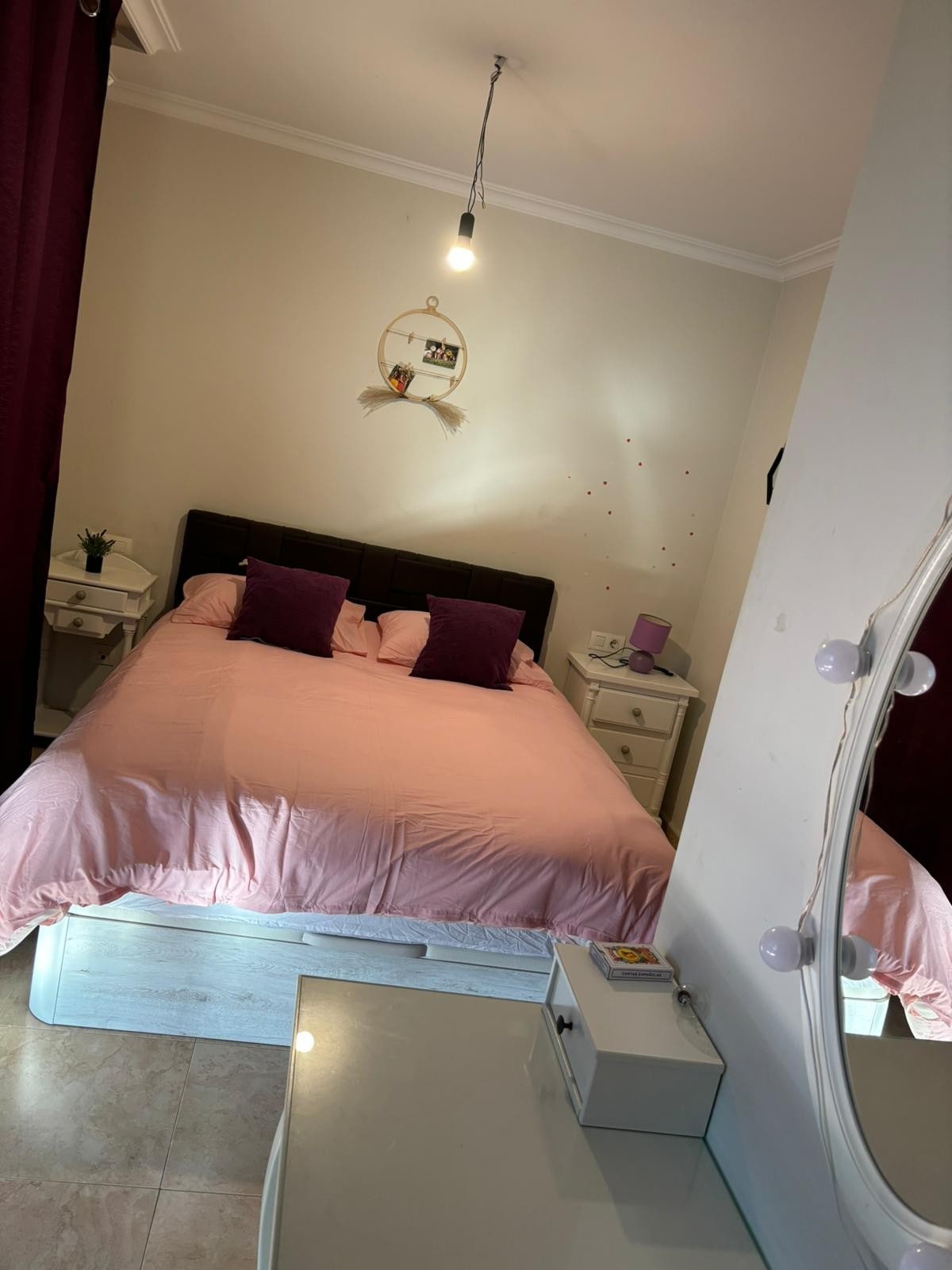 A well-lit sleeping area features a spacious bed with a soft pink coverlet and two decorative pillows. Flanking the bed are bedside tables with small plants. A mirror and a desk are positioned within the room. Soft light fixtures provide illumination, enhancing the serene ambiance.