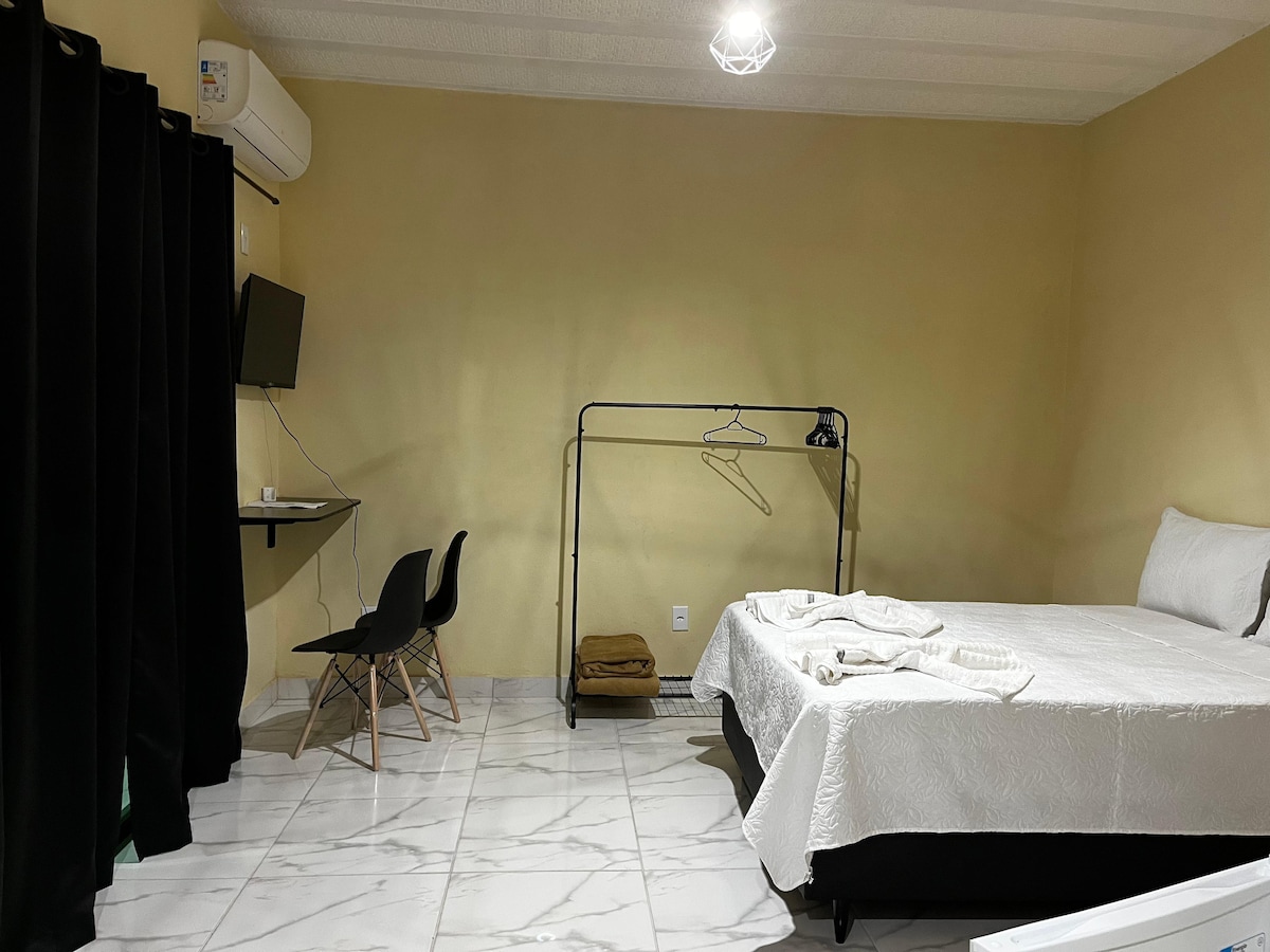 A well-lit room features a spacious bed with a white quilt. A small desk with two black chairs is positioned adjacent to the bed. A clothing rack stands nearby, with a television mounted on the wall. Dark curtains frame a window, enhancing privacy.