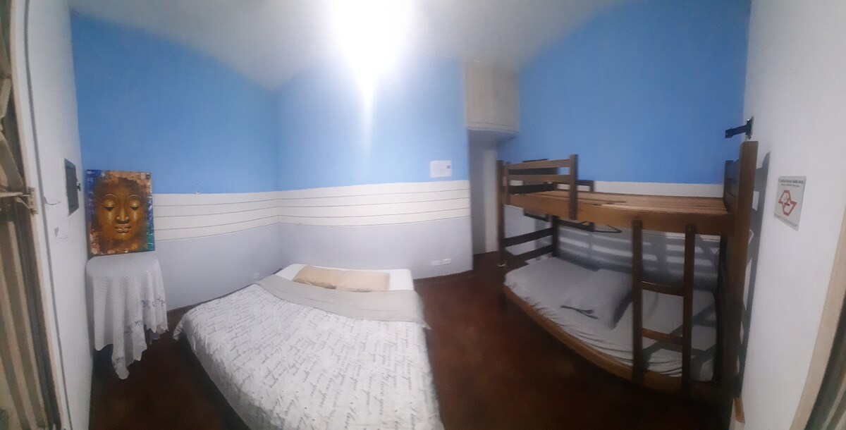 A brightly lit sleeping area features a spacious bed with light bedding, complemented by a wooden bunk bed in the corner. Light blue walls contribute to a serene atmosphere. A small table is positioned against the wall, enhancing the room's functionality.