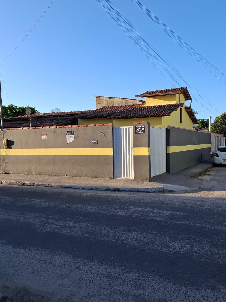 The exterior of a single-story house is shown, featuring a gray and yellow color scheme. A gated entrance is positioned on the side, and a white vehicle is parked nearby. The structure exhibits a tiled roof and a mix of wall materials.