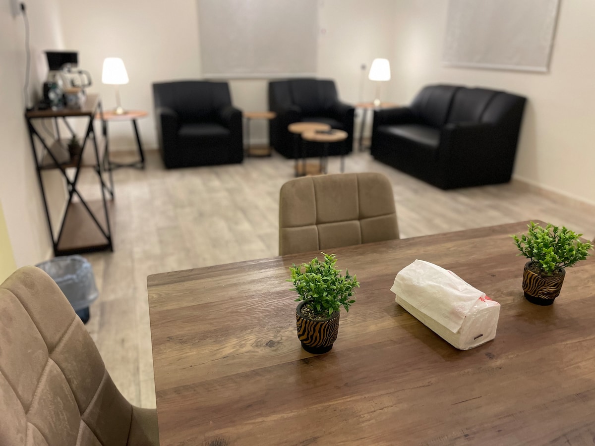 A spacious living area features two black sofas and a coffee table, along with soft lighting provided by two lamps. A dining table is visible in the foreground, adorned with small potted plants, contributing to the functional layout of the space.